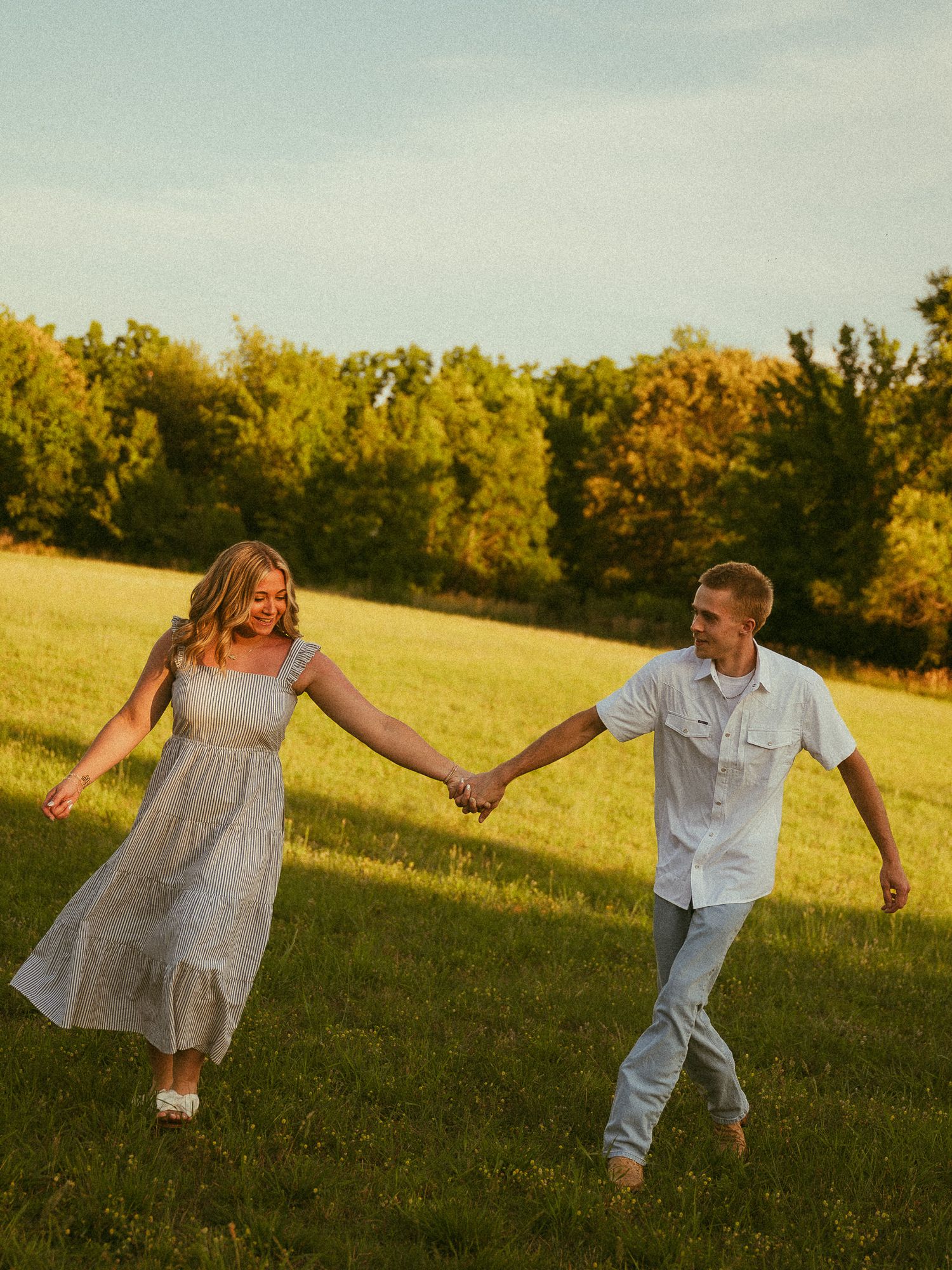 A couple holds hands and walks through a sunny green meadow surrounded by trees during golden hour.