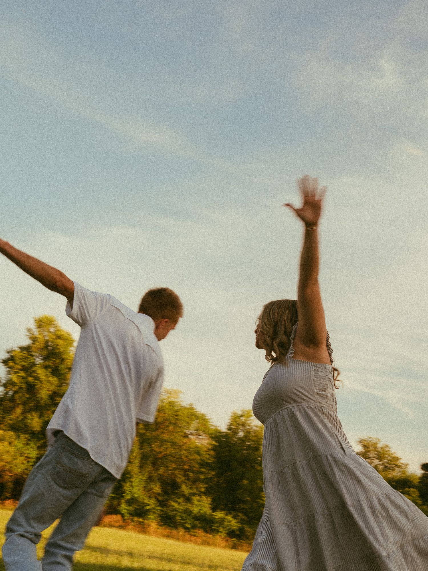 Two people dancing joyfully in a sunlit green field, arms raised, wearing casual summer clothes with trees in the background.