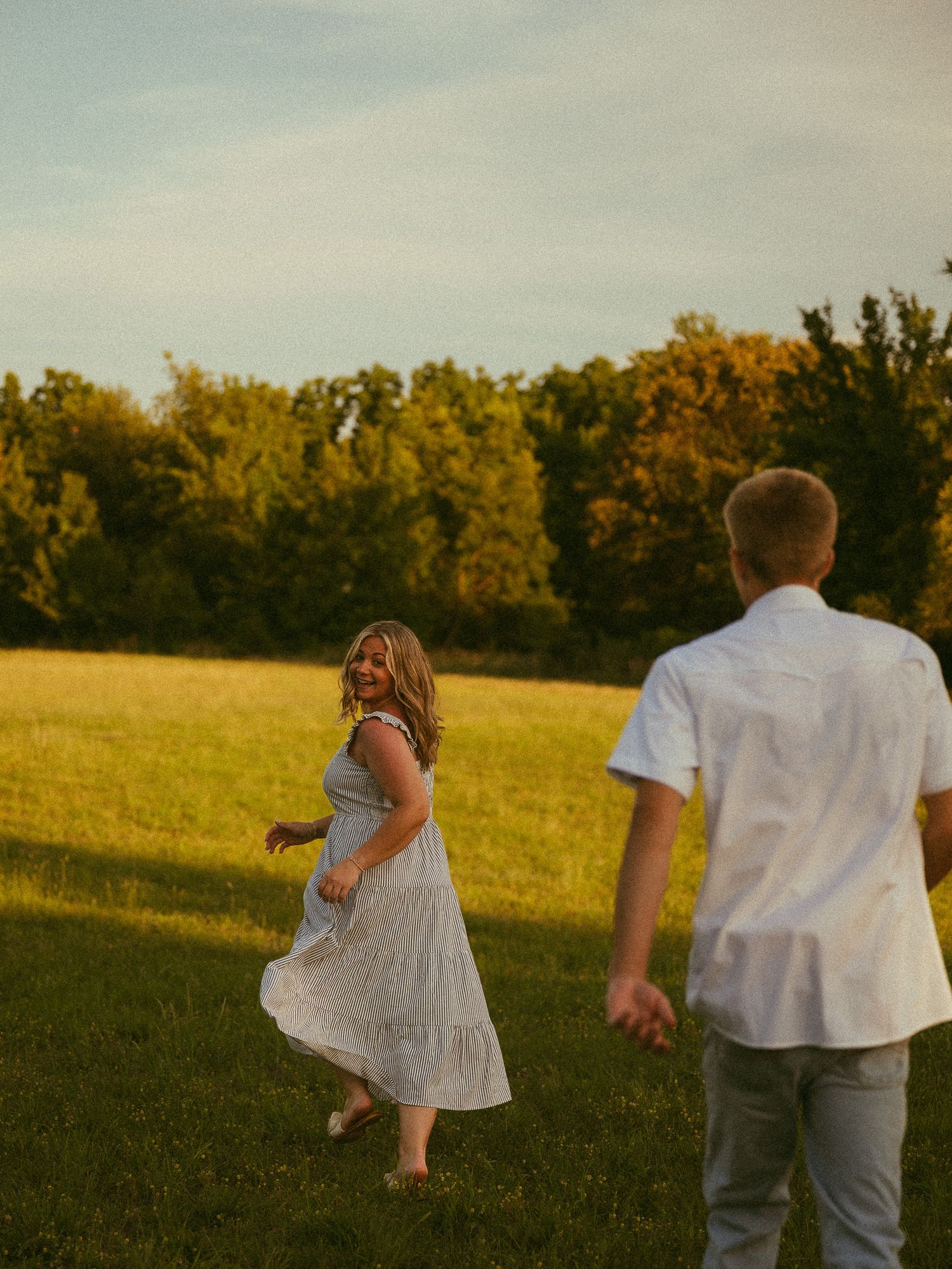 A couple enjoys a golden hour stroll in a lush green field, the woman in a white dress laughing joyfully.