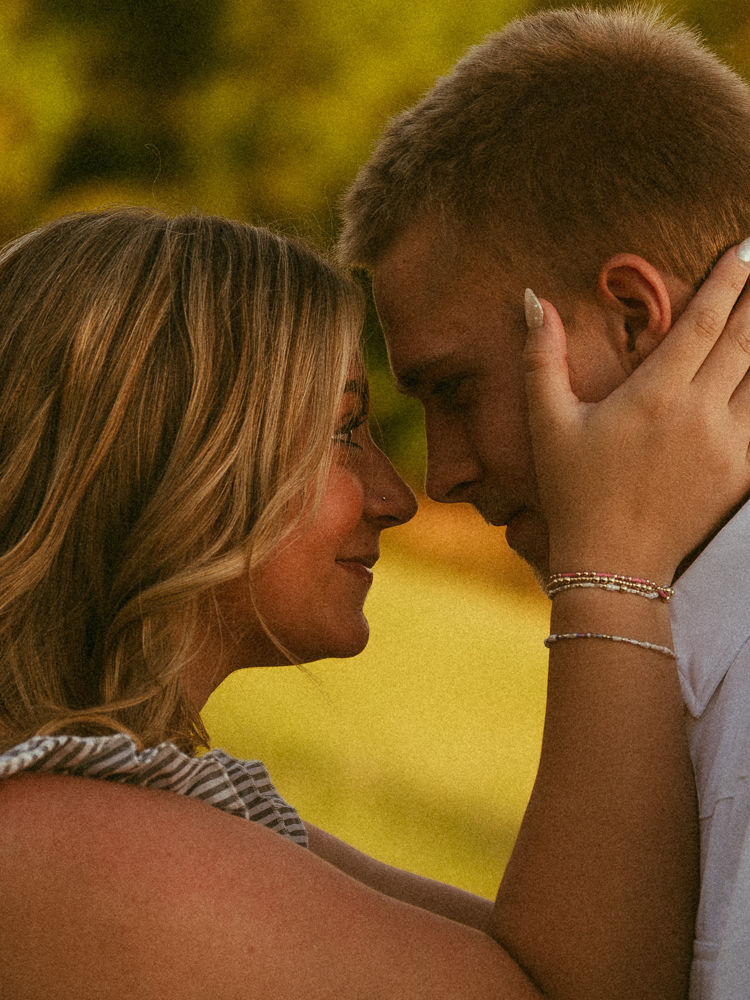 A couple shares a romantic forehead-to-forehead moment outdoors in warm golden hour light, the woman wearing a bracelet.