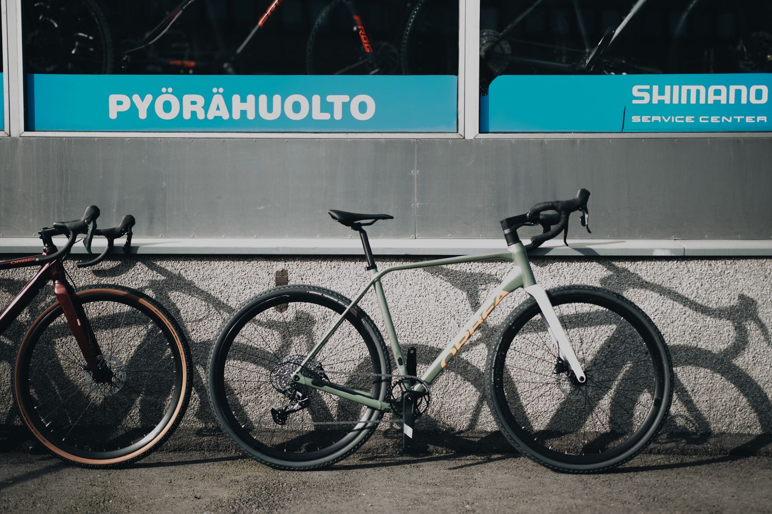 A white road bike parked against a wall with Shimano and Pyorahuolto signage, casting shadows on the surface.