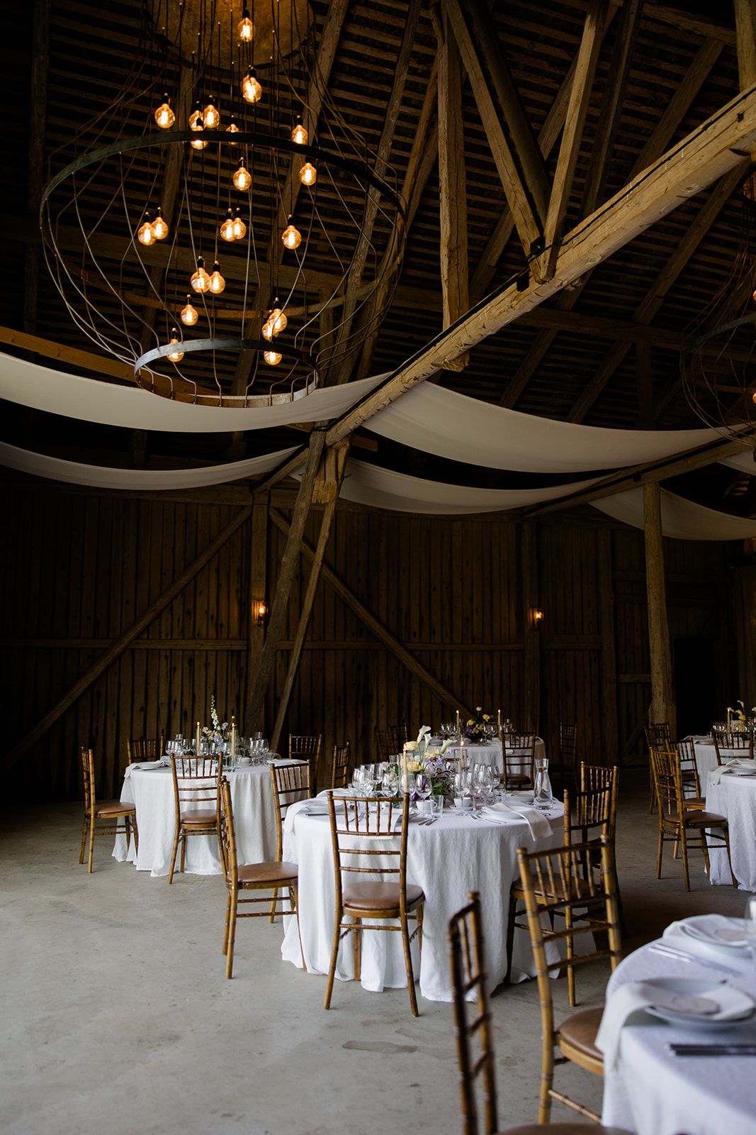 Rustic barn wedding reception setup with hanging Edison bulbs and wooden chiavari chairs around white-clothed tables.