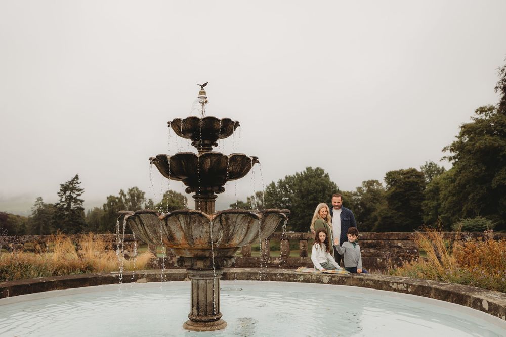 A couple sits on the edge of an ornate stone fountain in a misty garden setting with autumn foliage.