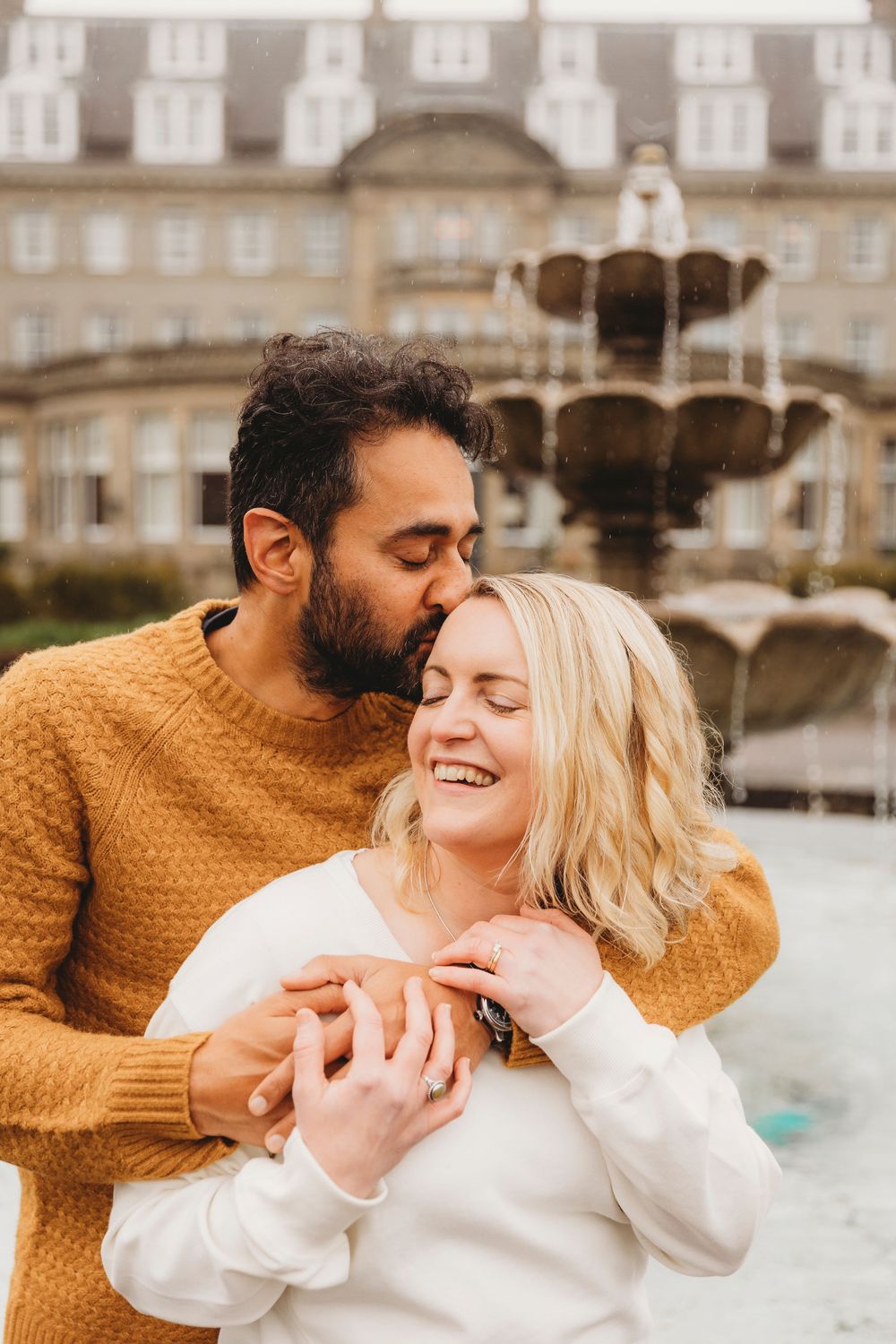 A romantic couple embraces and laughs together in front of an ornate fountain and historic building exterior.