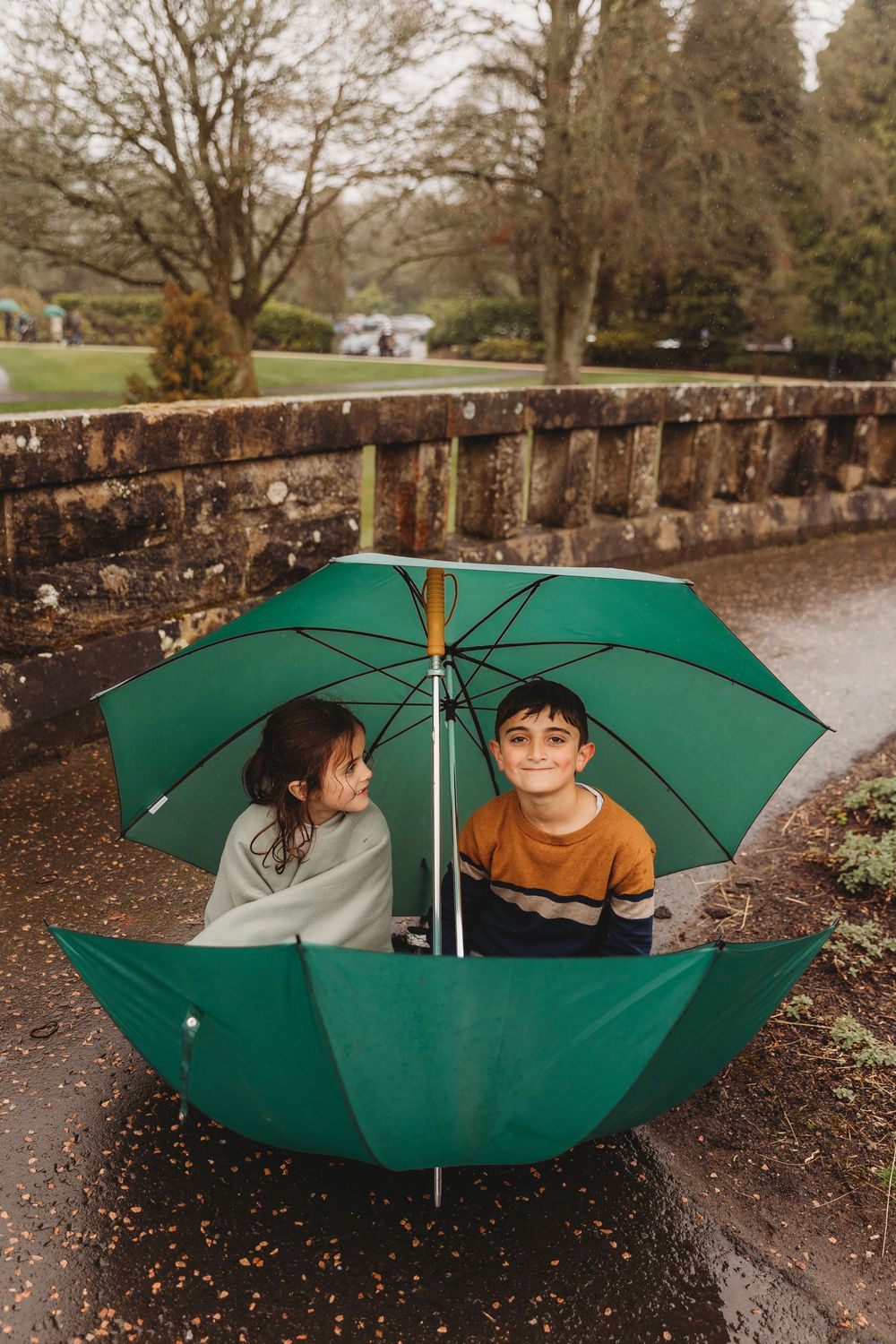 A sequence of photos showing two people playfully posing with a large green umbrella in a park setting on a rainy day.