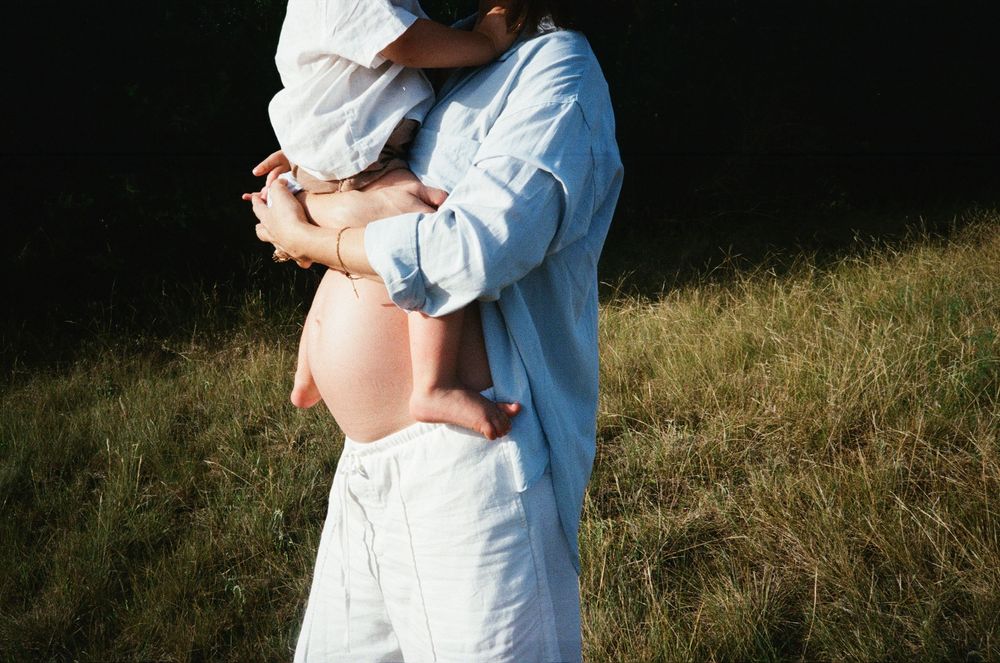 A tender maternity moment captured in soft sunlight against a grassy field backdrop.