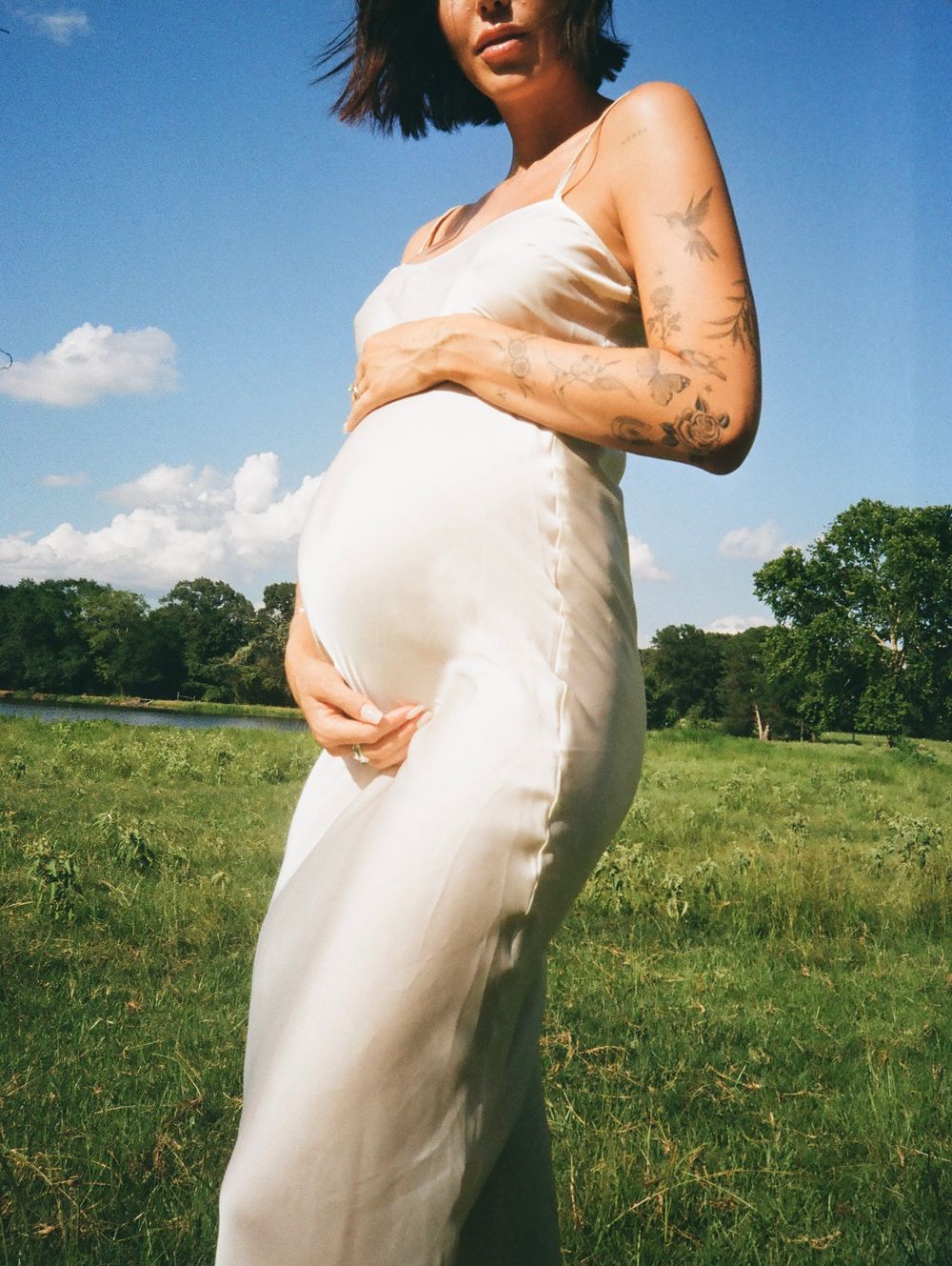 Maternity photos in white dress against sunny meadow landscape.