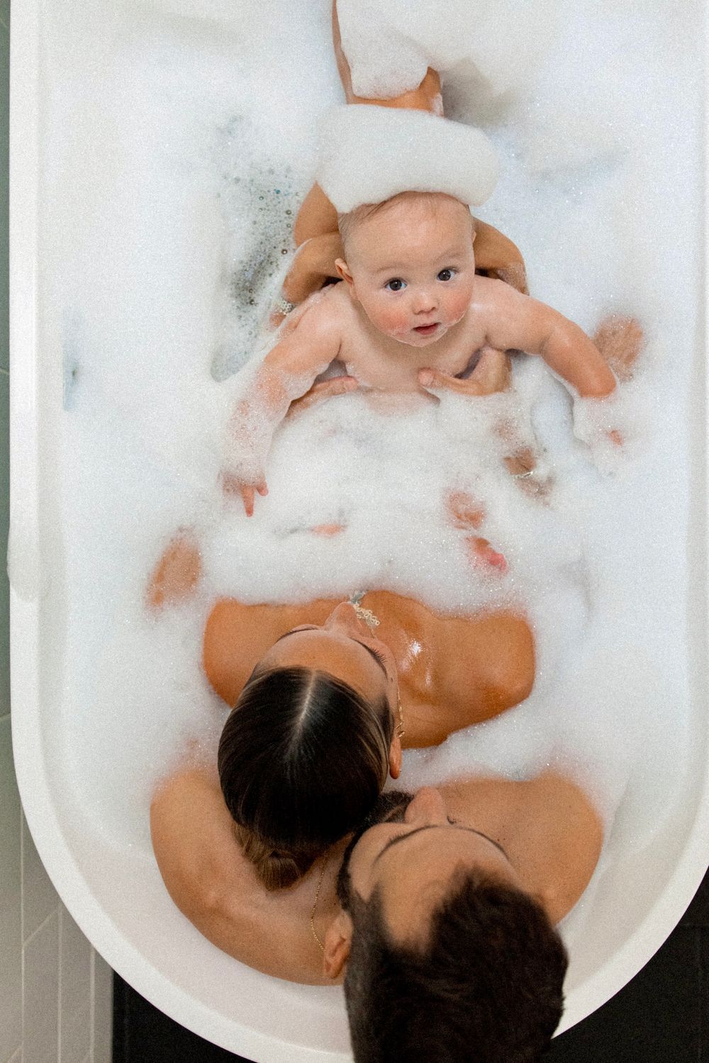 Baby enjoying bath time in white tub filled with bubbles and soapy foam.