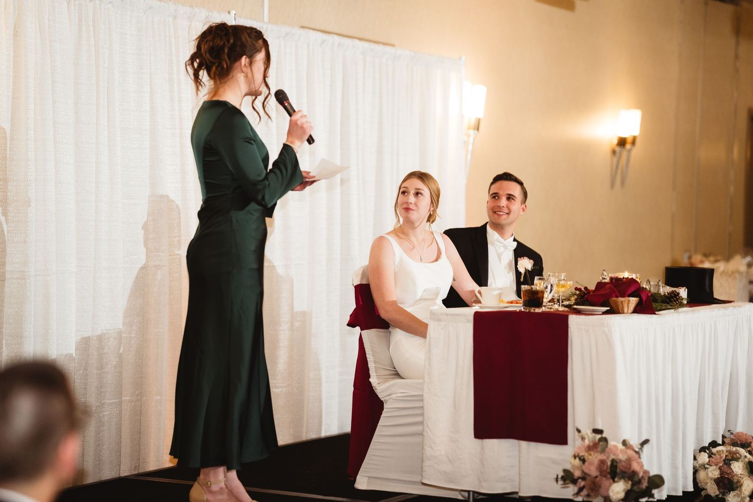 A speaker in a dark green dress delivers a speech at an elegant wedding reception with burgundy and white decor.