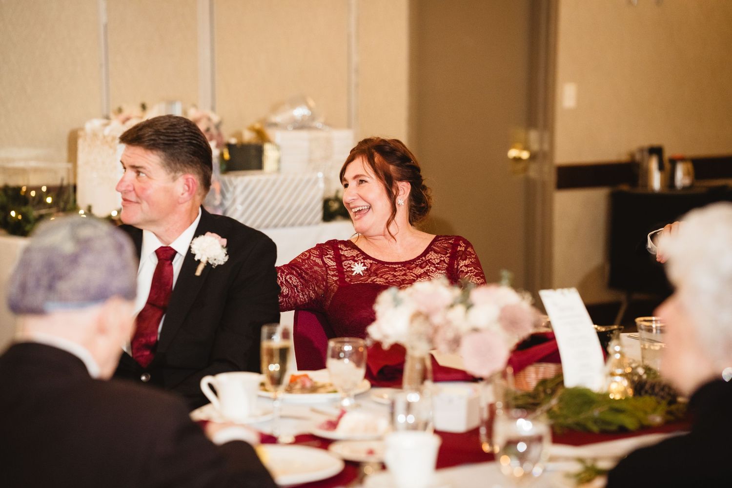Wedding reception guests seated at long tables with burgundy accents and floral centerpieces enjoy dinner and conversation.