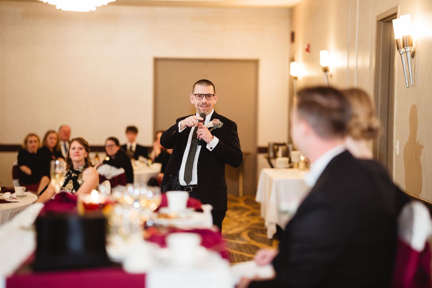 A wedding reception speech being delivered in an elegant banquet room with burgundy and white table settings.