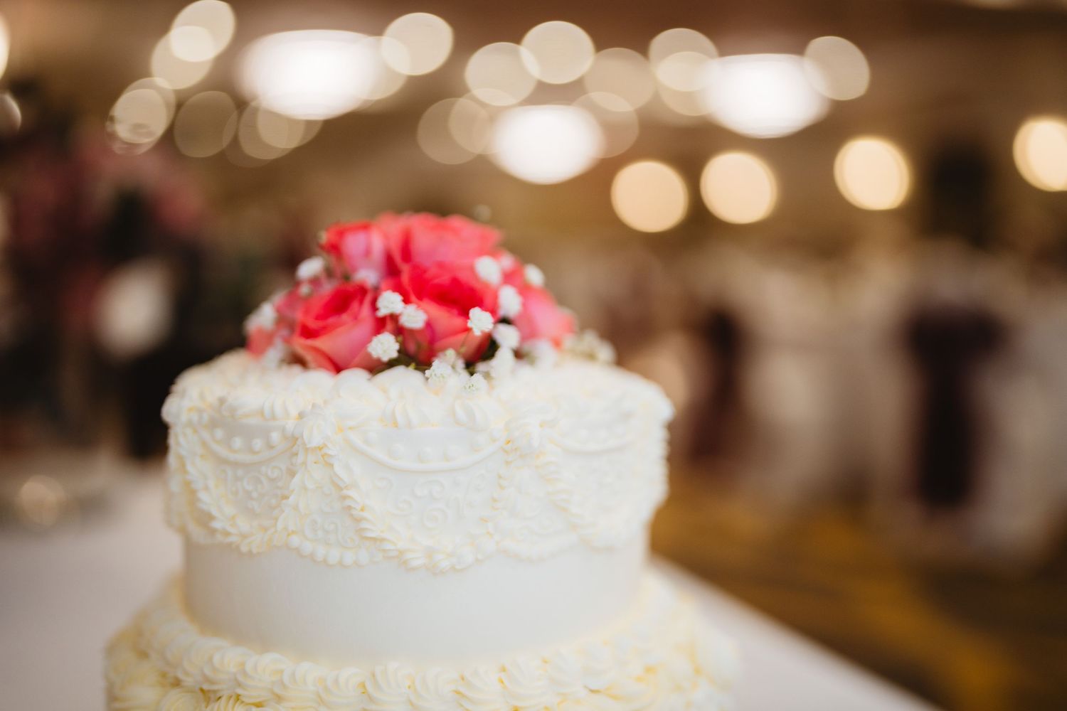 A white wedding cake decorated with red flowers sits elegantly on display with soft bokeh lights in the background.