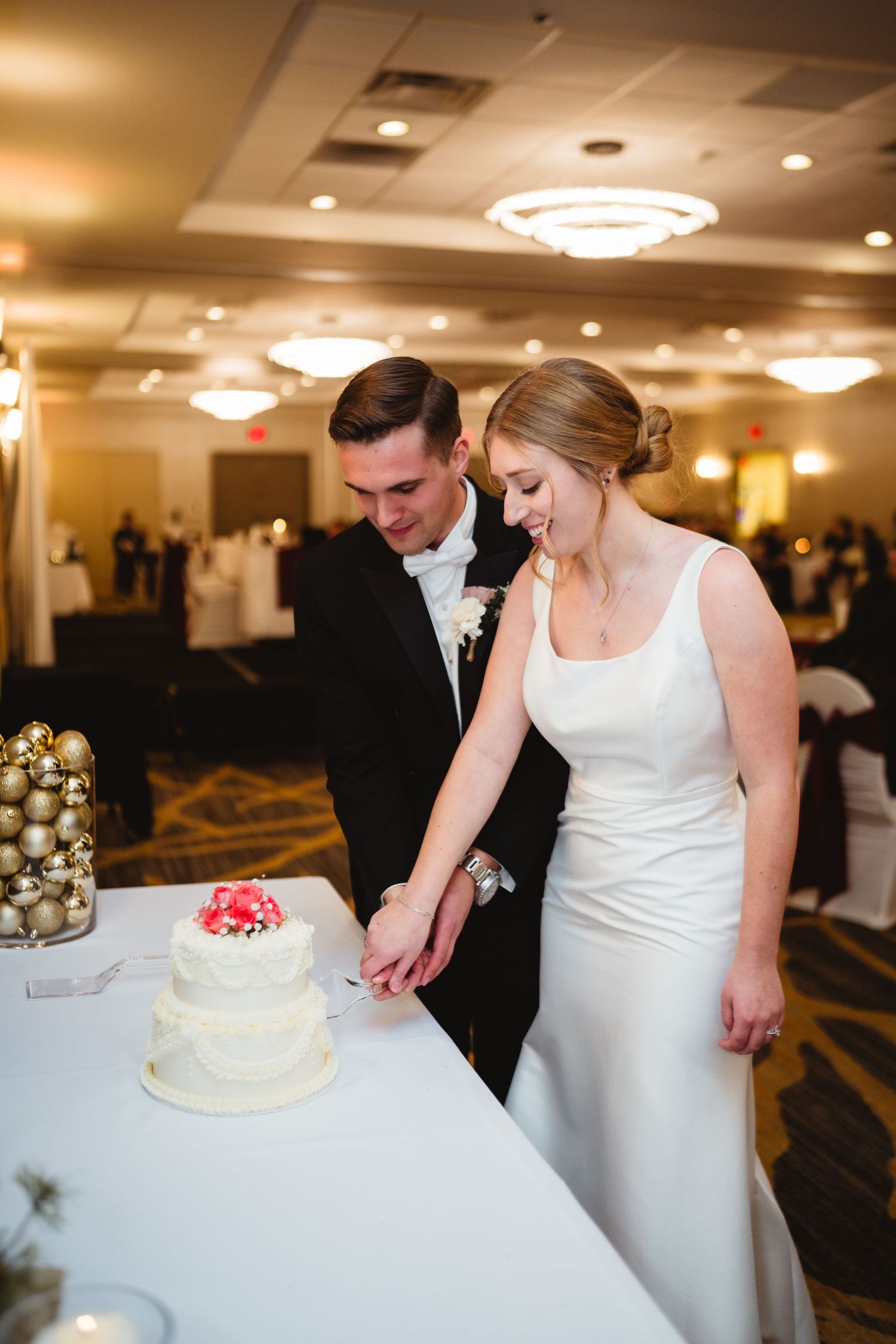 A couple in formal wedding attire cuts into their white wedding cake topped with red berries at an elegant reception venue.