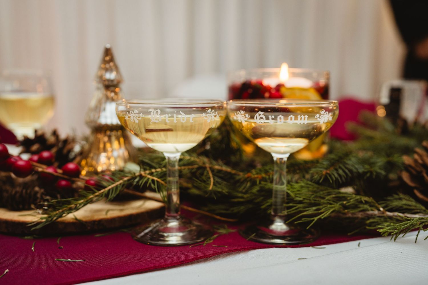 Two vintage champagne coupes with sparkling wine on a burgundy tablecloth with evergreen branches and holiday decor.