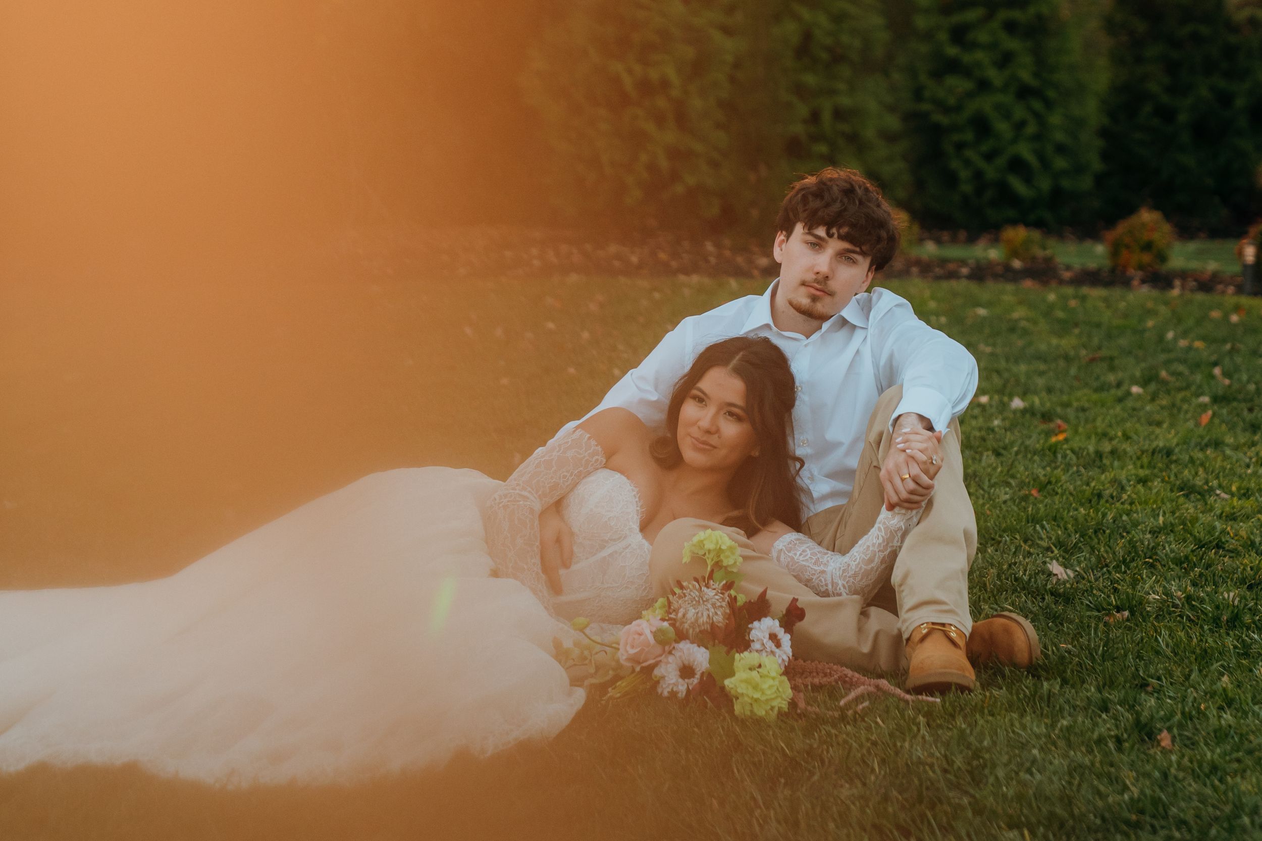 A couple in wedding attire lies together on grass during sunset creating a romantic outdoor portrait.