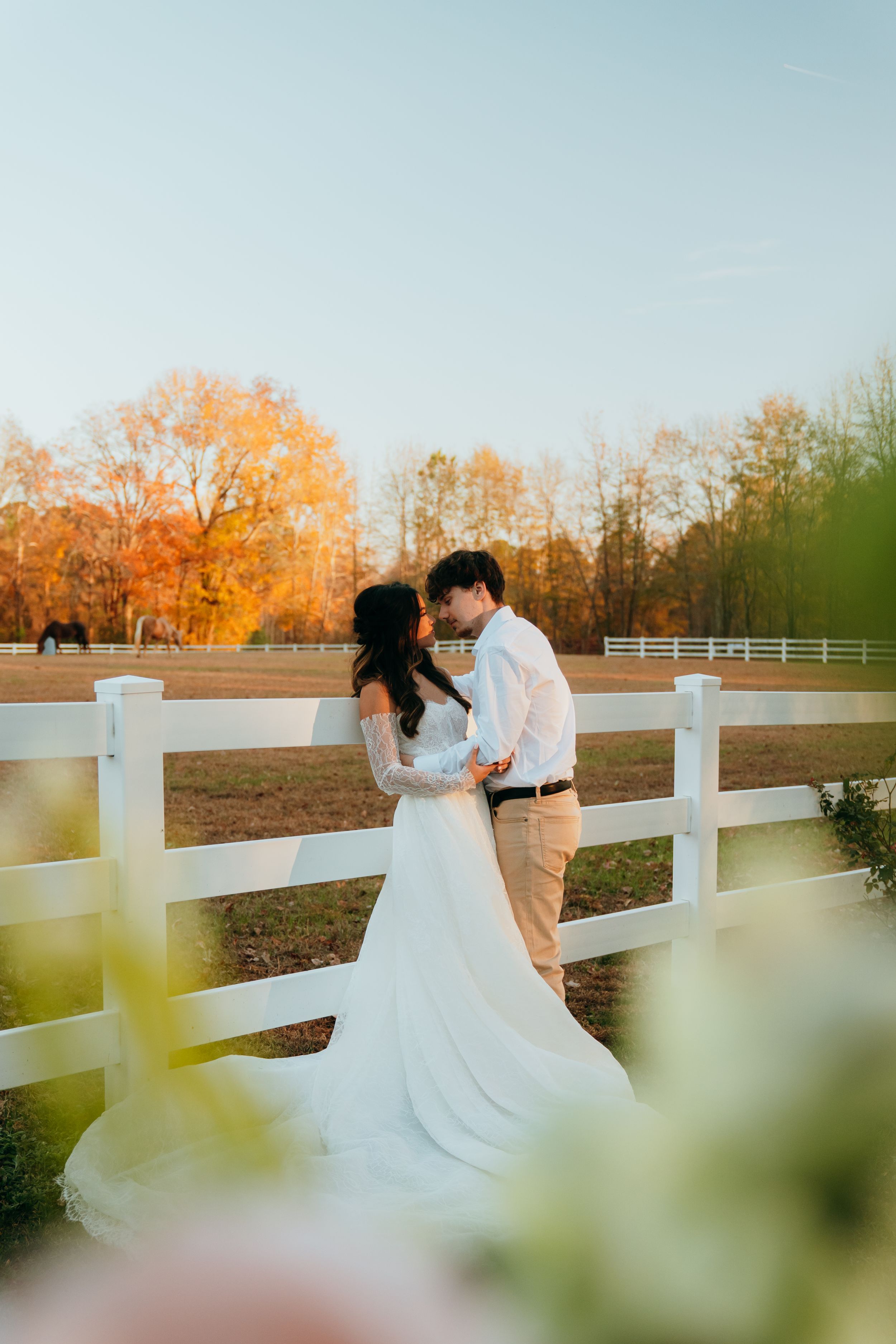 A romantic sunset wedding photoshoot along a white fence with autumn trees glowing orange in the background.