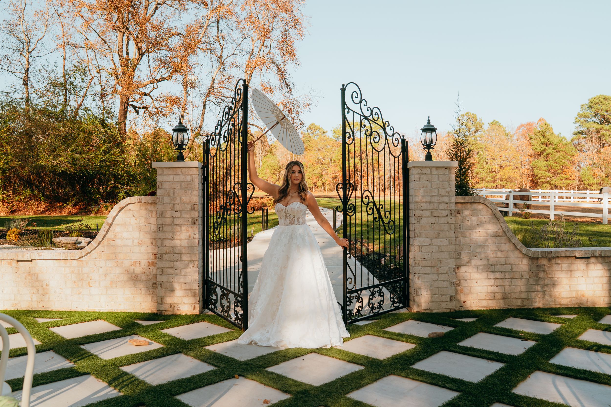 A white wedding gown stands elegantly in front of ornate black iron gates at a garden entrance during autumn.