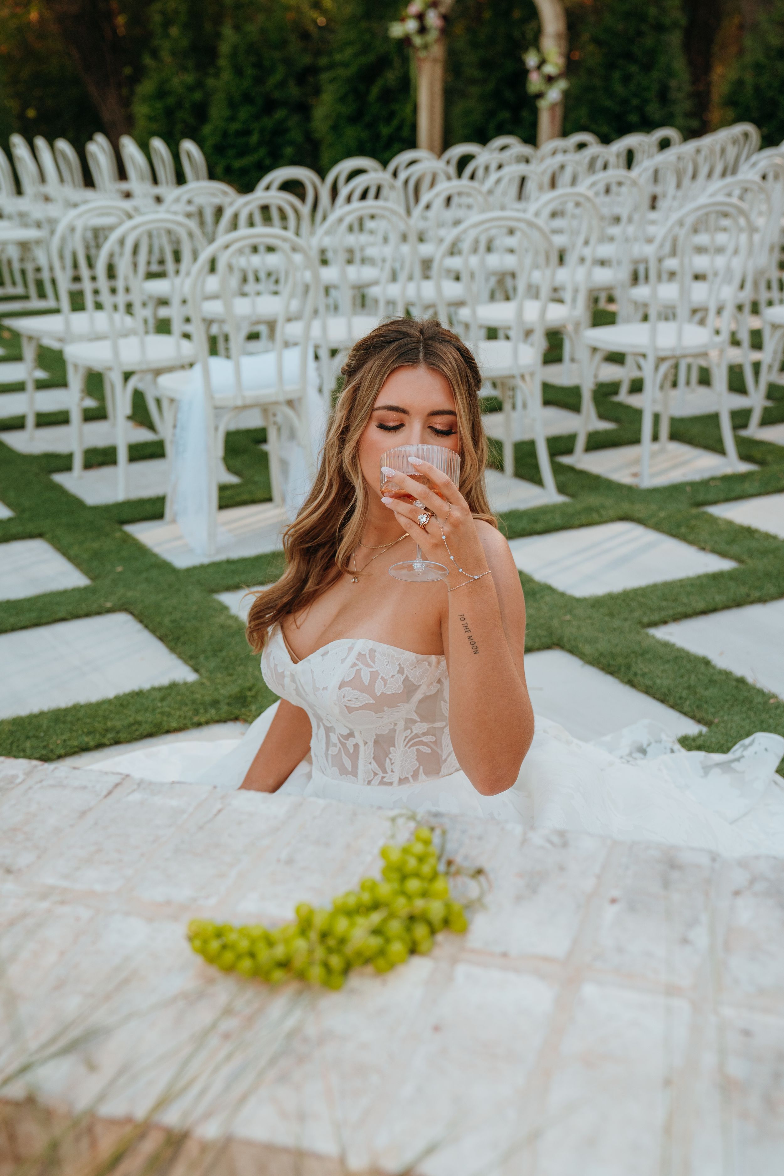 A bride in a white lace corset gown sits at an outdoor wedding venue with rows of white chairs and fresh grapes on the table.