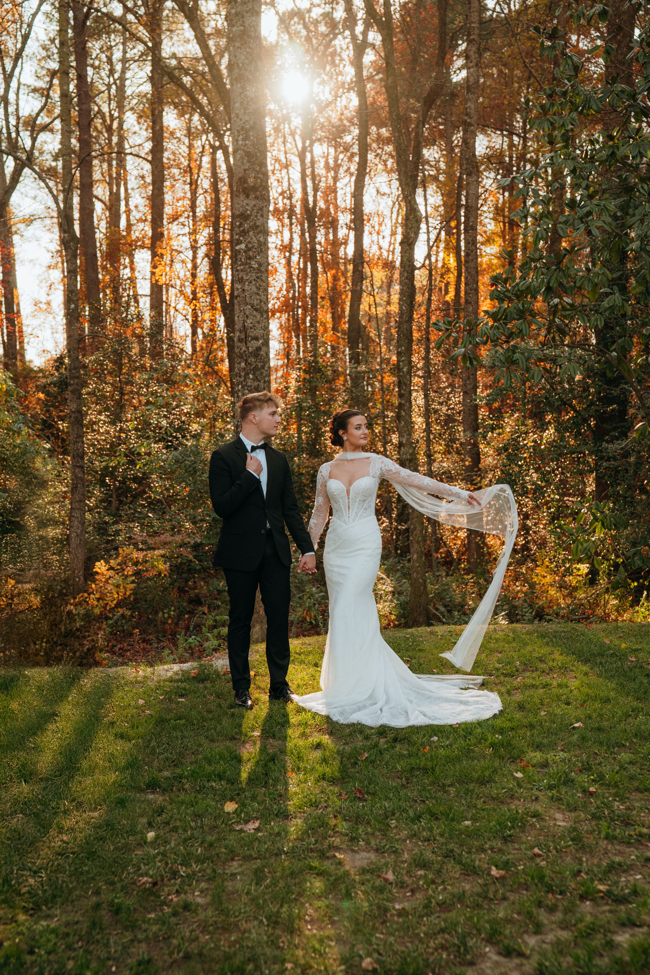 A couple poses in a sun-drenched forest during golden hour as a wedding veil flows gracefully in the autumn breeze.