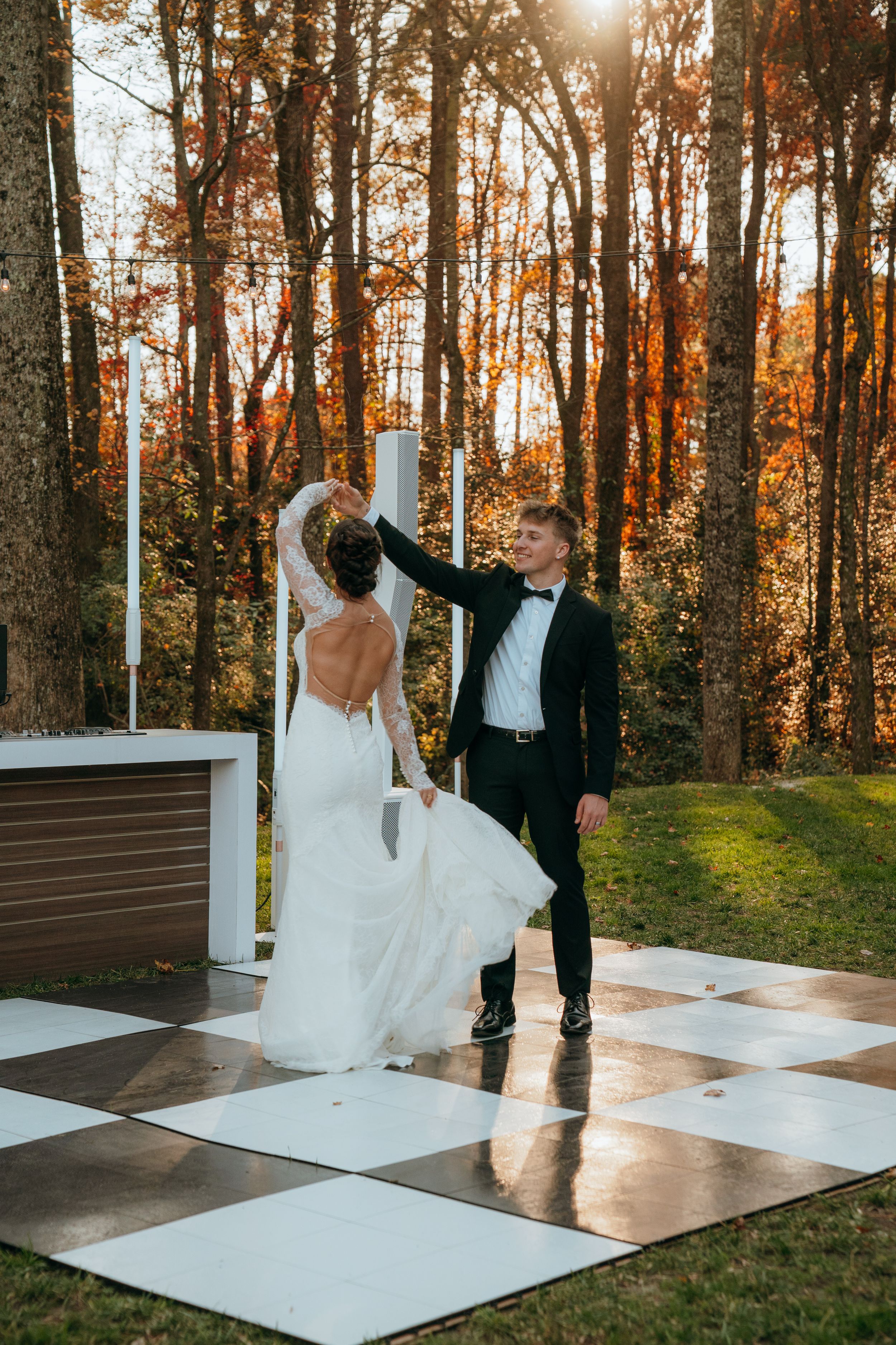 A couple dances together on an outdoor dance floor at sunset with autumn trees in the background.
