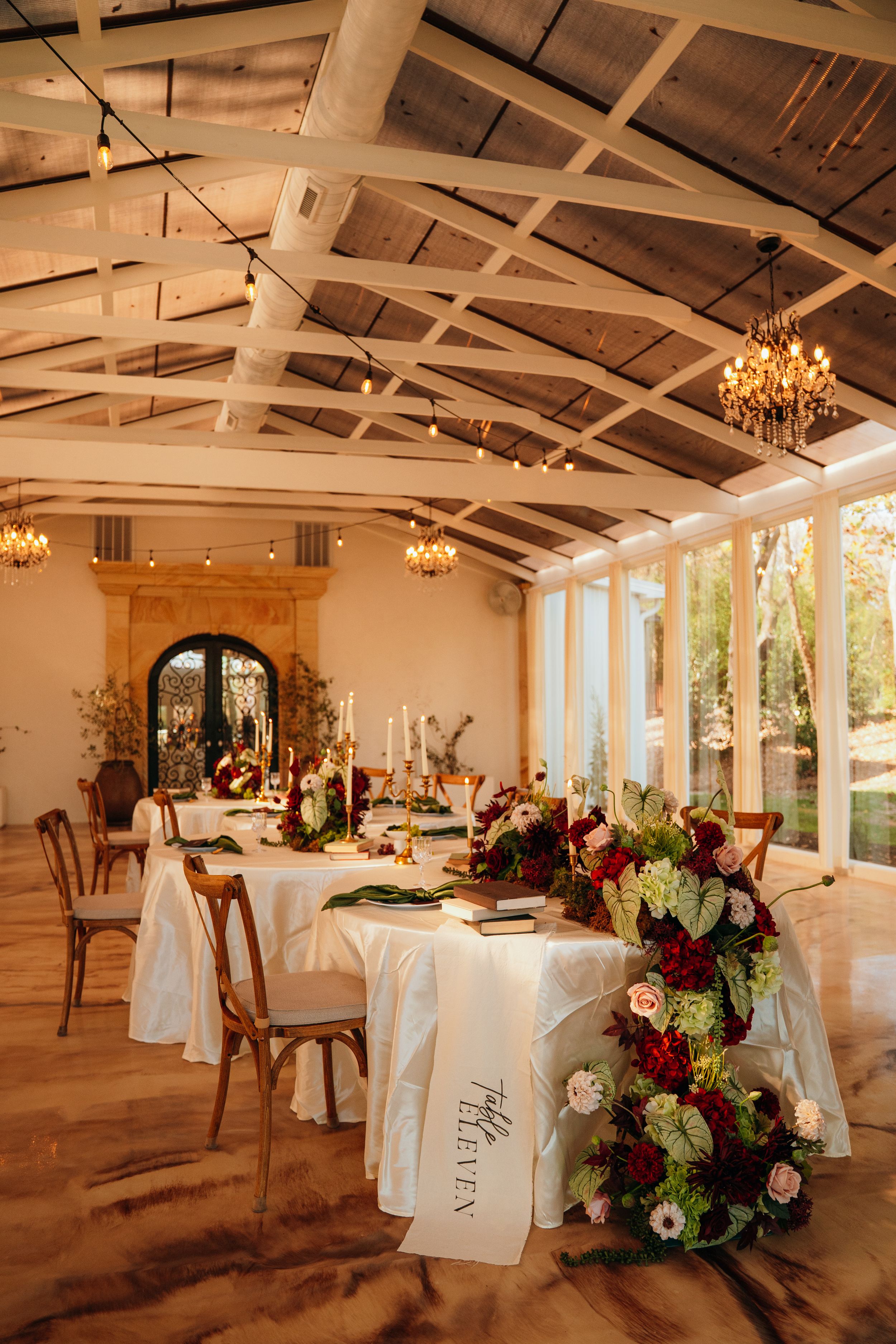 Elegant wedding reception venue with rustic wood beams, crystal chandeliers, and white-clothed tables with floral centerpieces.