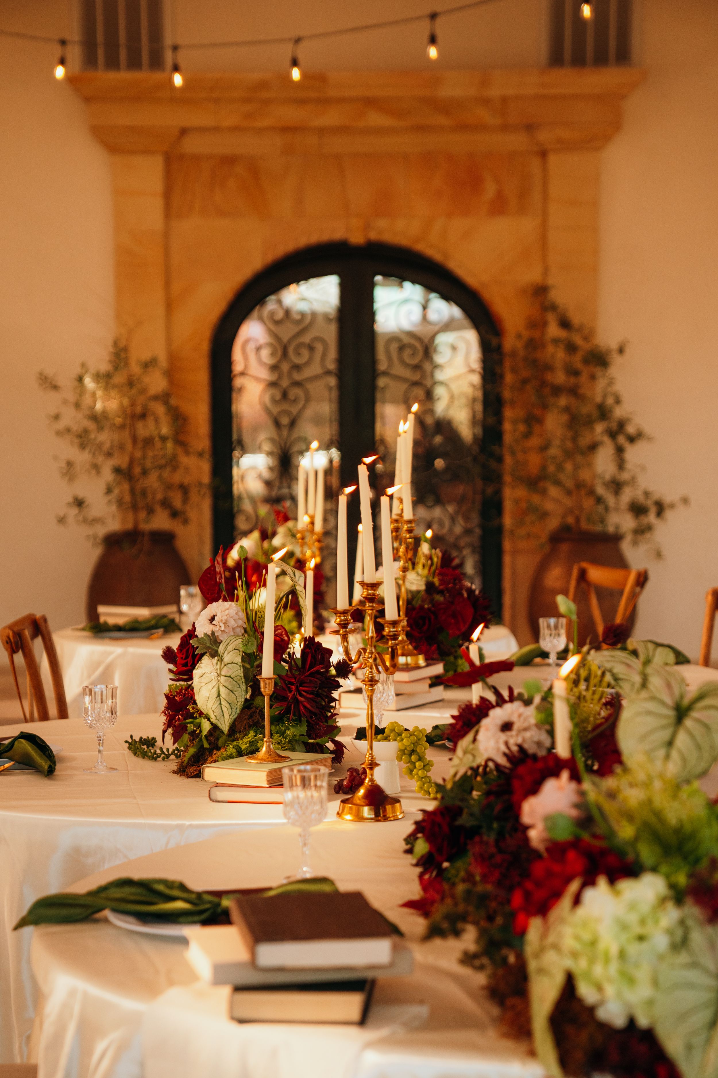 Elegant wedding table setting with tall white candles and burgundy floral arrangements against an arched window backdrop.