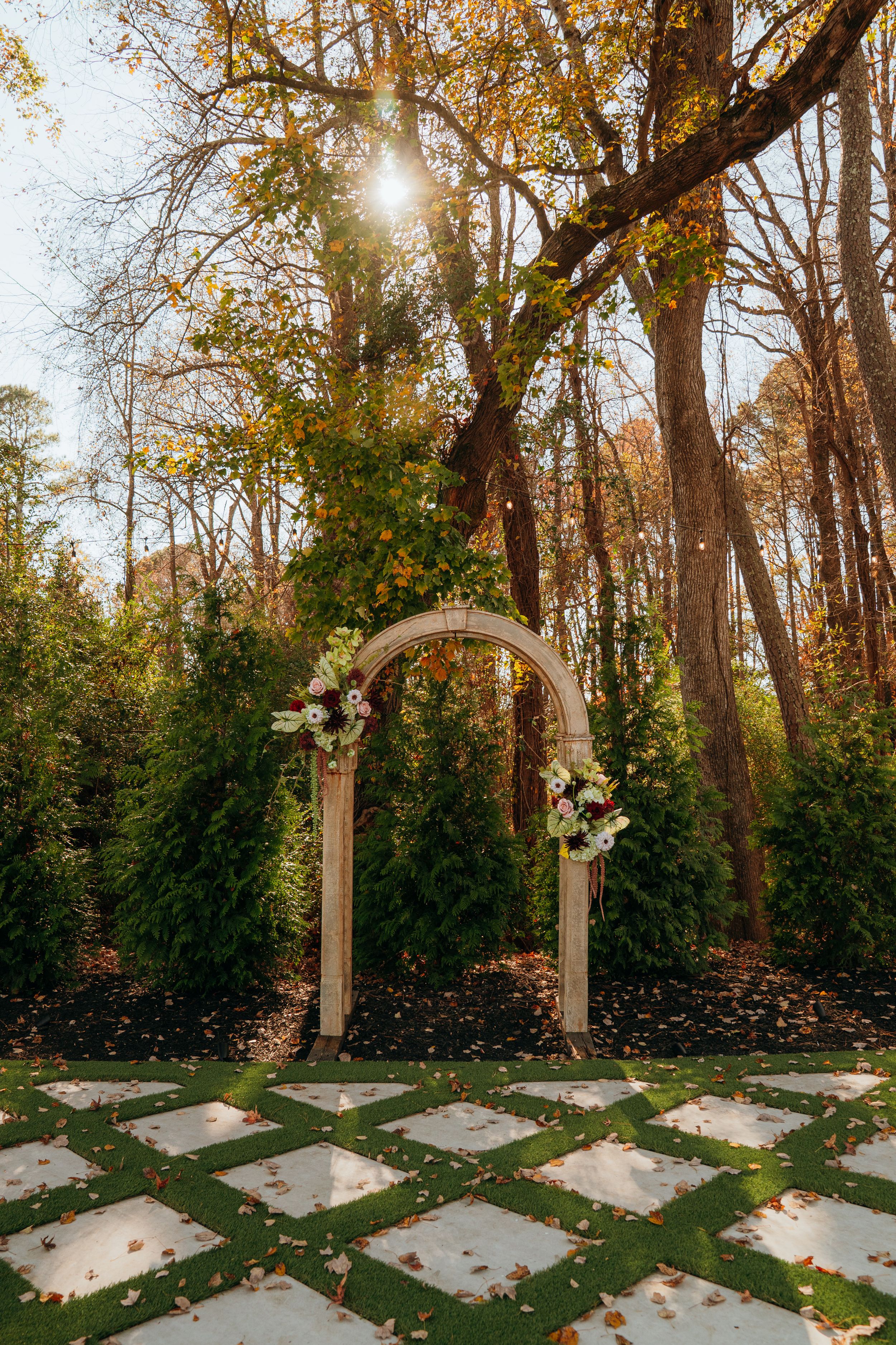 A white wooden wedding arch decorated with flowers stands in a forest setting with stone pavers and autumn foliage.