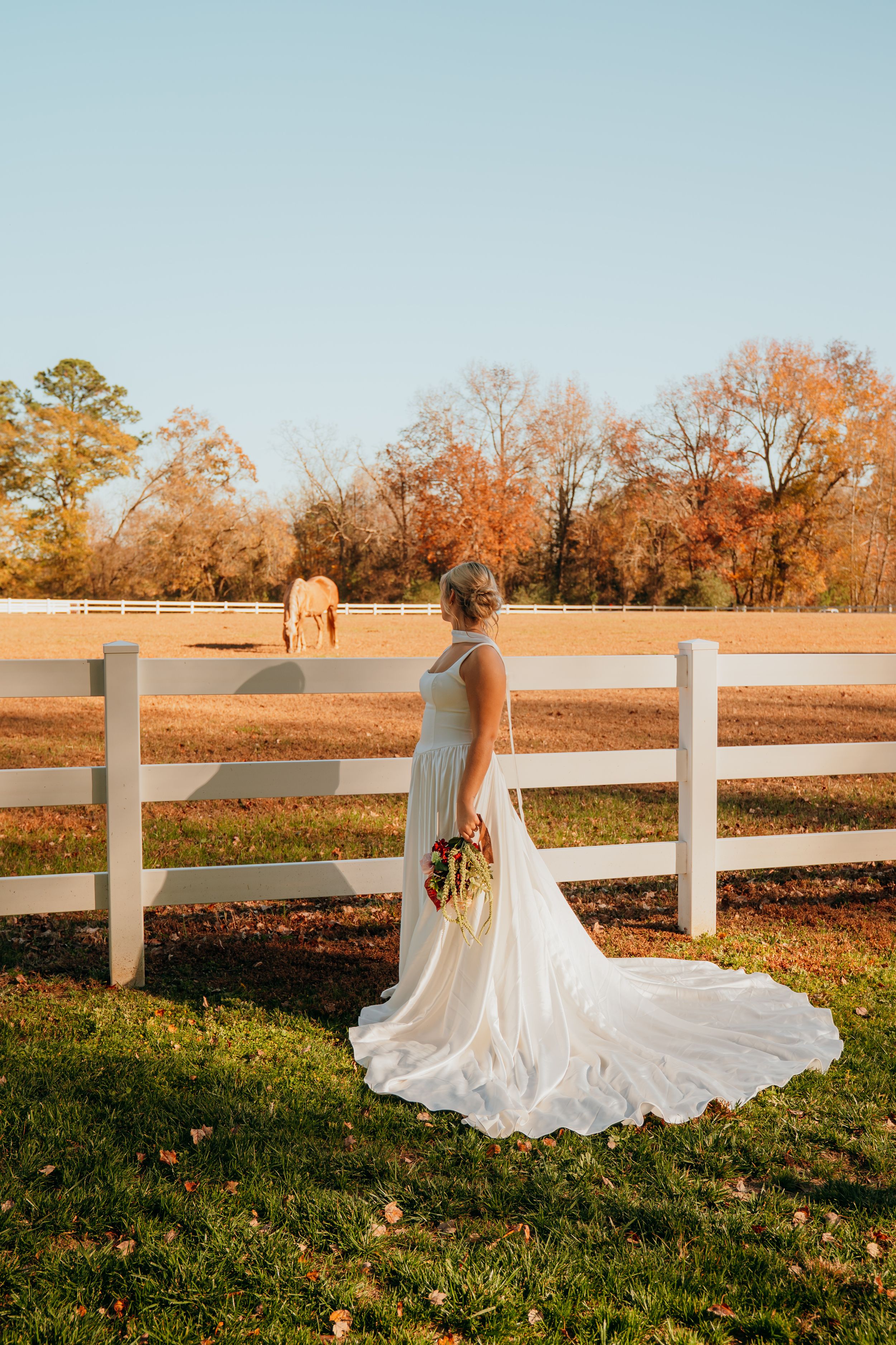Elegant white wedding gown with long train flows across autumn grass beside white fence at sunset.