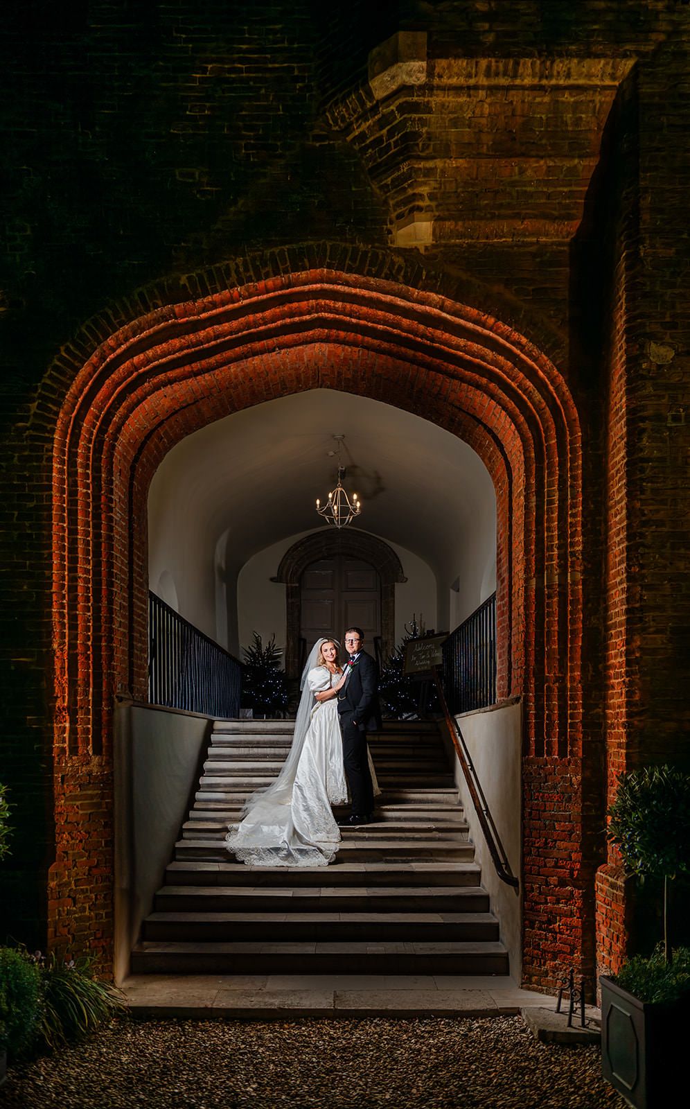A wedding couple stands in a dramatic brick archway with stairs and moody lighting creating a romantic atmosphere.