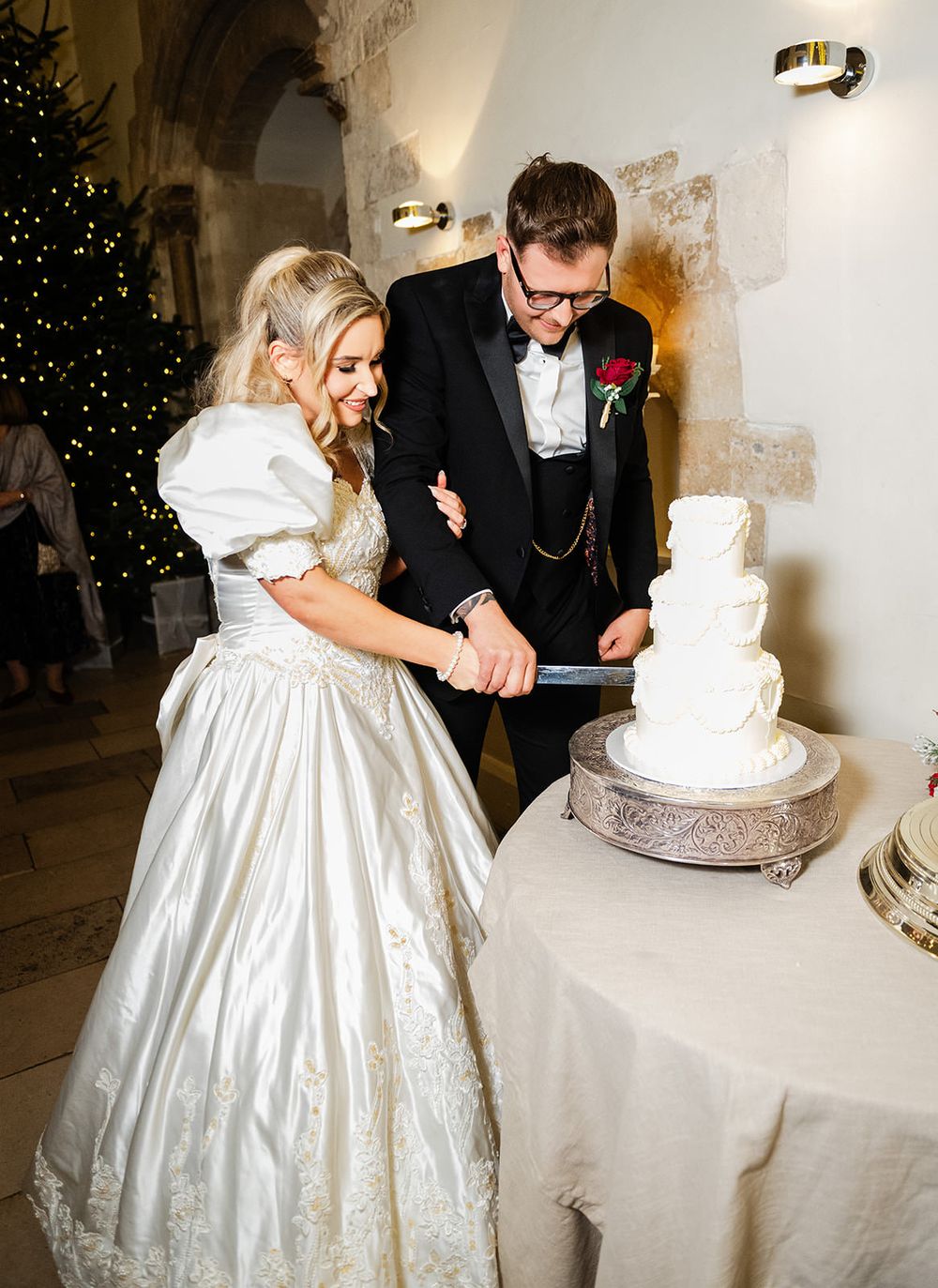 A couple in formal wedding attire cuts their elegant white tiered wedding cake together at their reception.