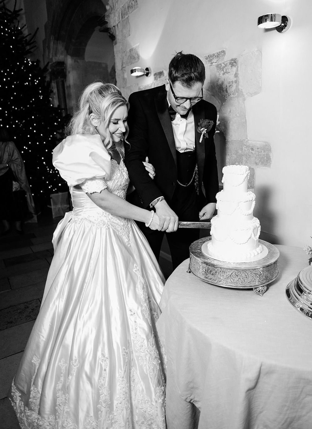 A couple in formal wedding attire cuts their elegant white tiered wedding cake at their reception.