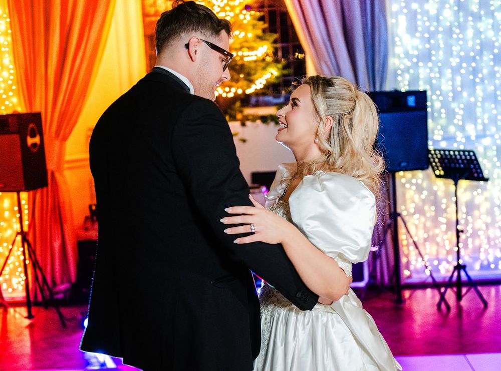 A couple shares their first dance at a wedding reception with colorful orange and purple drapery in the background.
