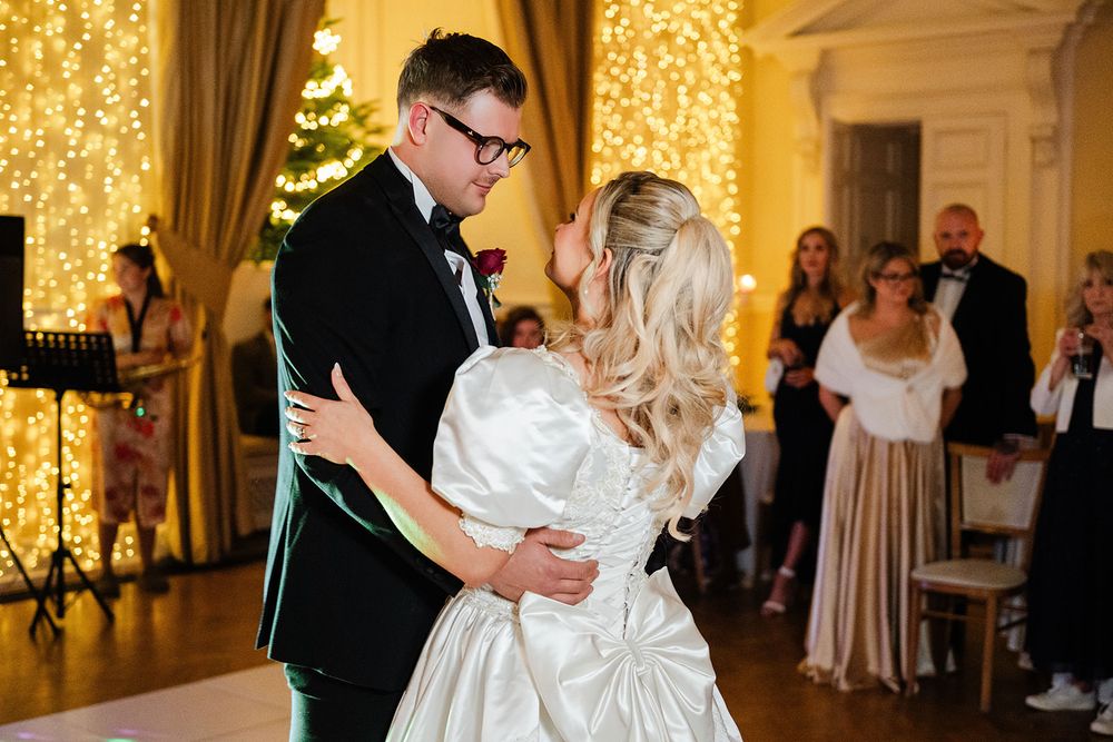A wedding couple shares their first dance in an elegant ballroom with warm glowing lights and onlooking guests.