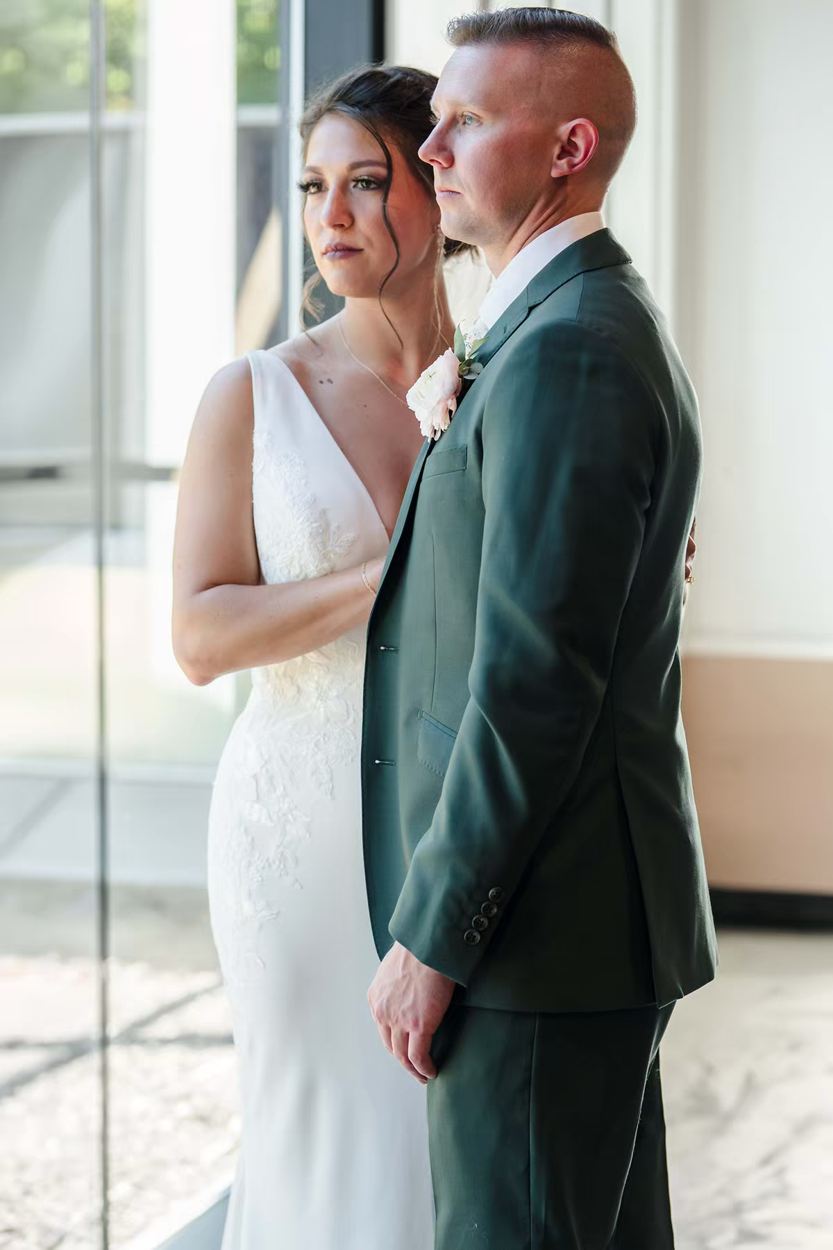 A couple in wedding attire, with the bride in white and groom in emerald green suit, pose together near a window in Retreat 21 Wedding Venue