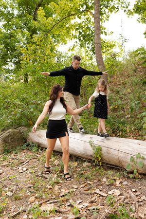 Couple playfully balances on fallen log in wooded area during outdoor photoshoot.