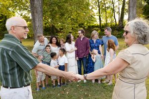 Group of people dancing in a circle outdoors under green trees during a summer gathering.