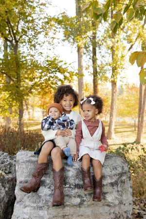 Three children in cowboy boots sit together on a large rock during autumn with golden foliage in the background.