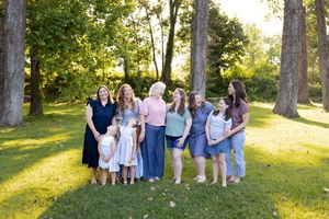A group of people in light summer dresses laughing and walking together in a sunlit park with tall trees.
