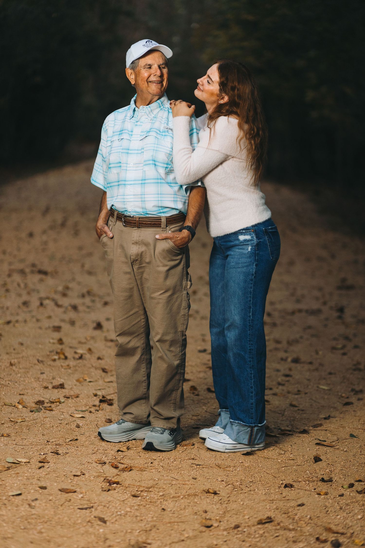 Two people stand together on a dirt path wearing casual clothing in a warm outdoor setting.