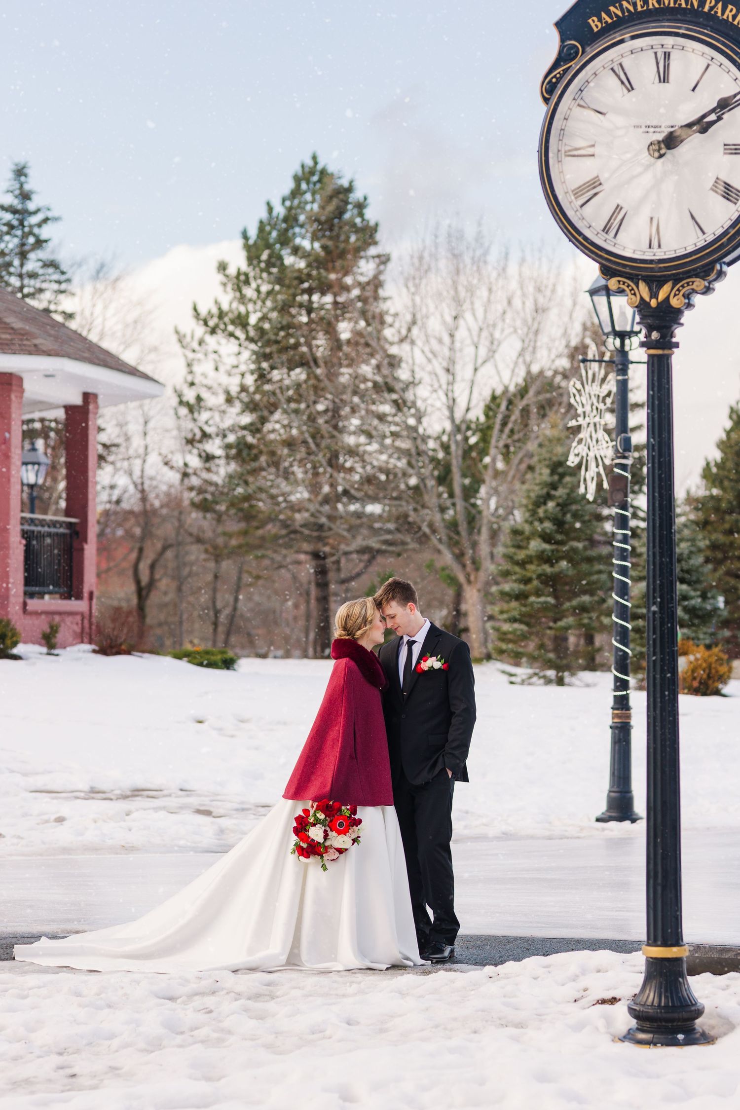 A couple in winter wedding attire poses near a vintage clock post in a snowy landscape.