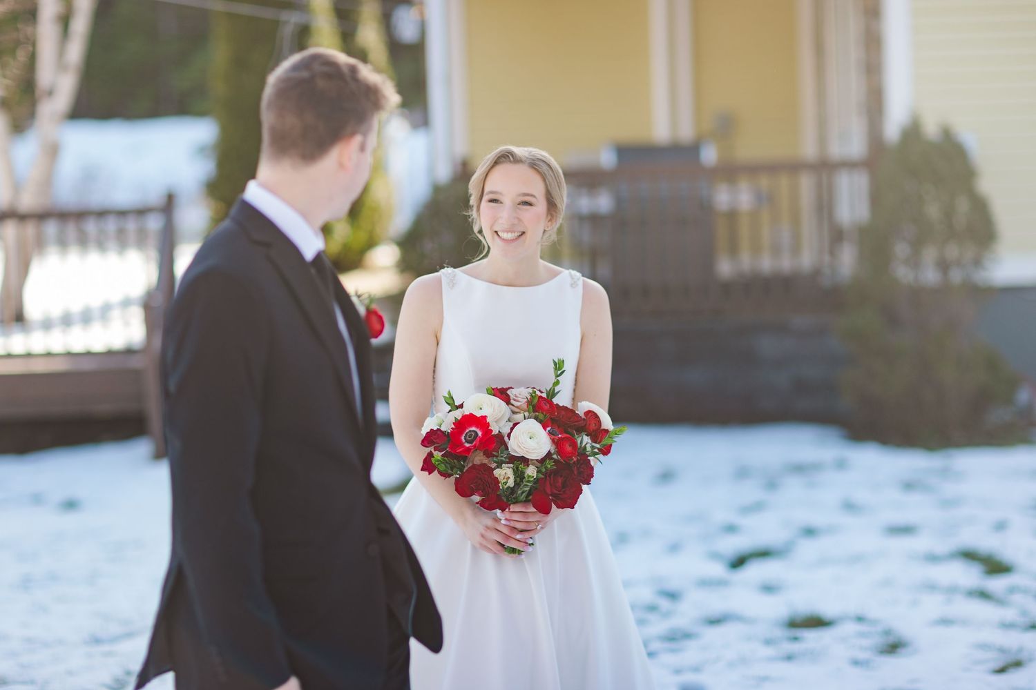 Wedding party poses in front of a yellow house on a snowy winter day with red bouquets.