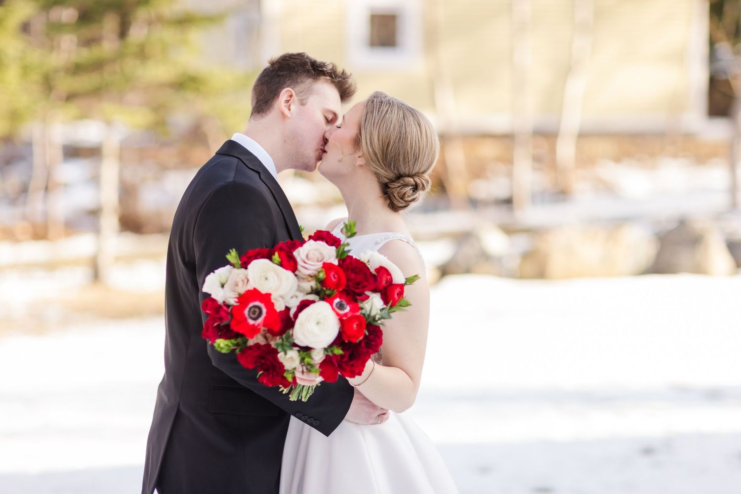 A couple shares a romantic kiss in winter while holding a vibrant red and white bouquet against a snowy backdrop.