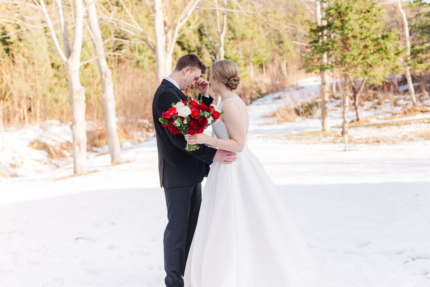 A couple in formal wedding attire embraces in a snowy winter landscape with evergreen trees in the background.
