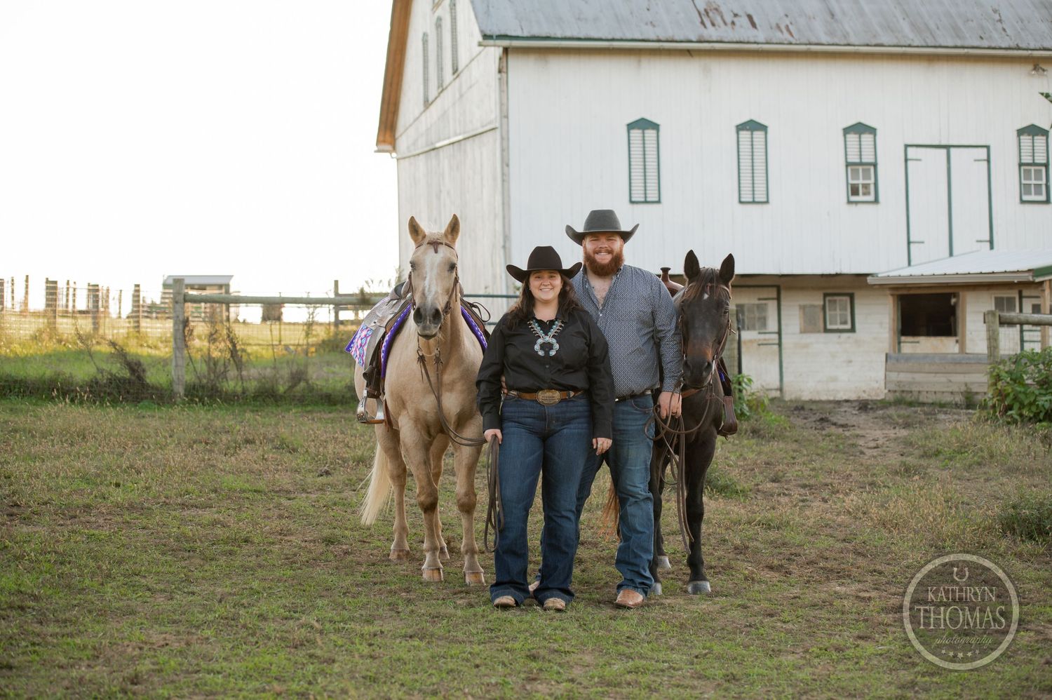 Two people with horses pose in front of white barn at dusk.