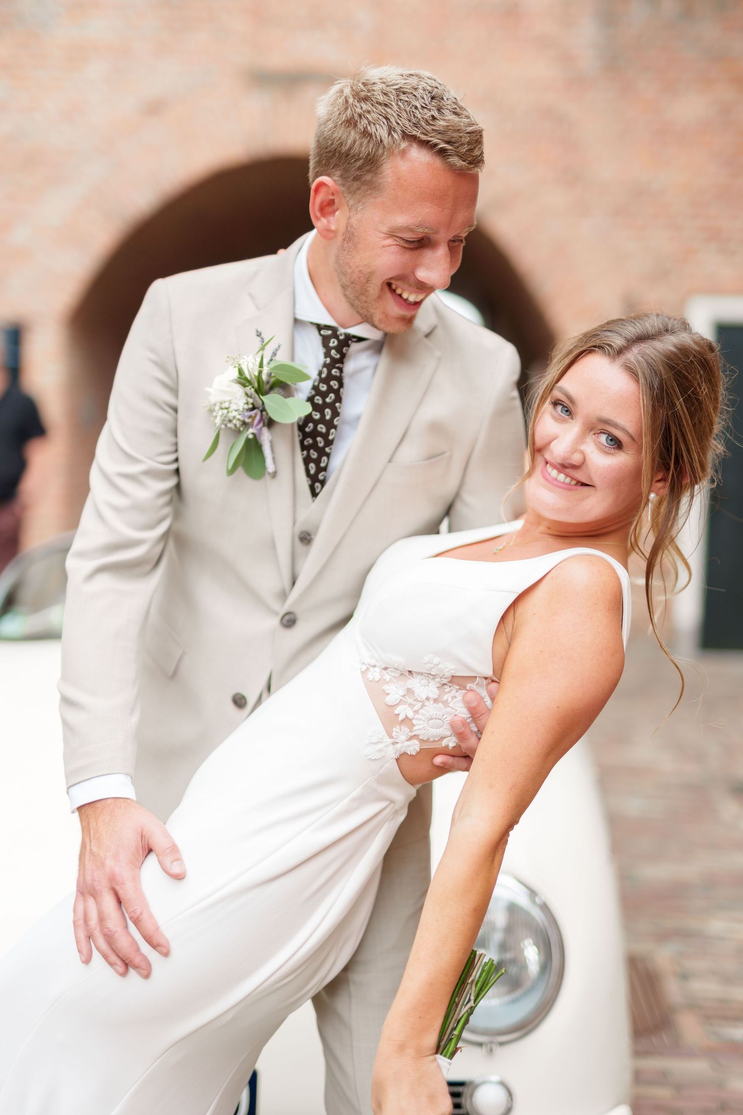 A happy couple shares a joyful moment on their wedding day in front of a vintage white car and brick archway.
