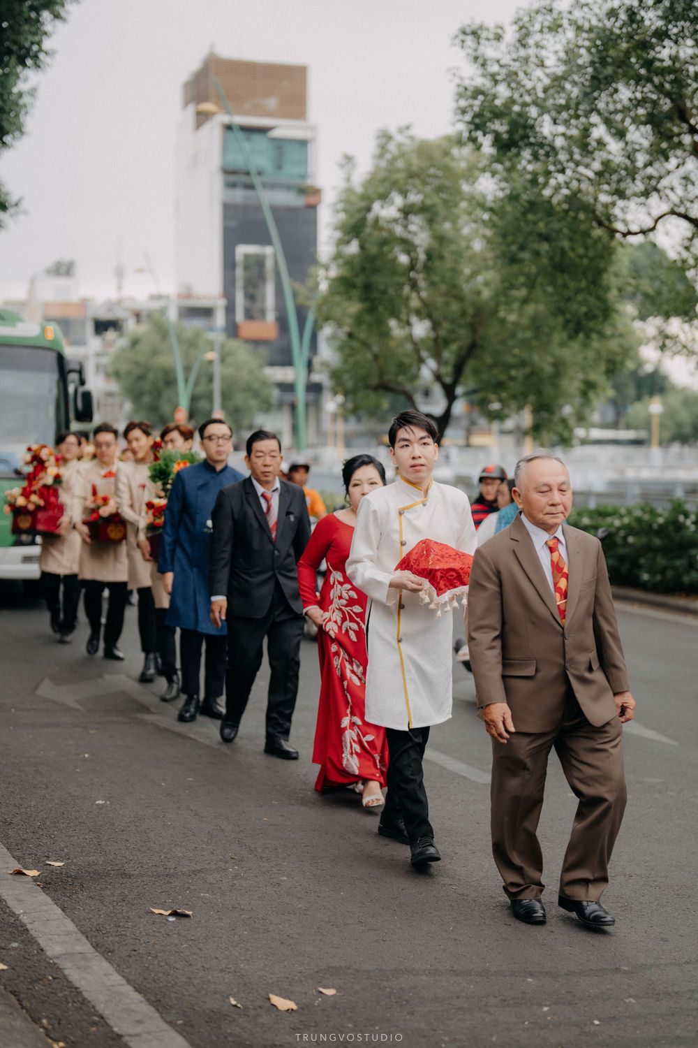 Groom in white ao dai leads traditional Vietnamese wedding procession down a tree-lined street.