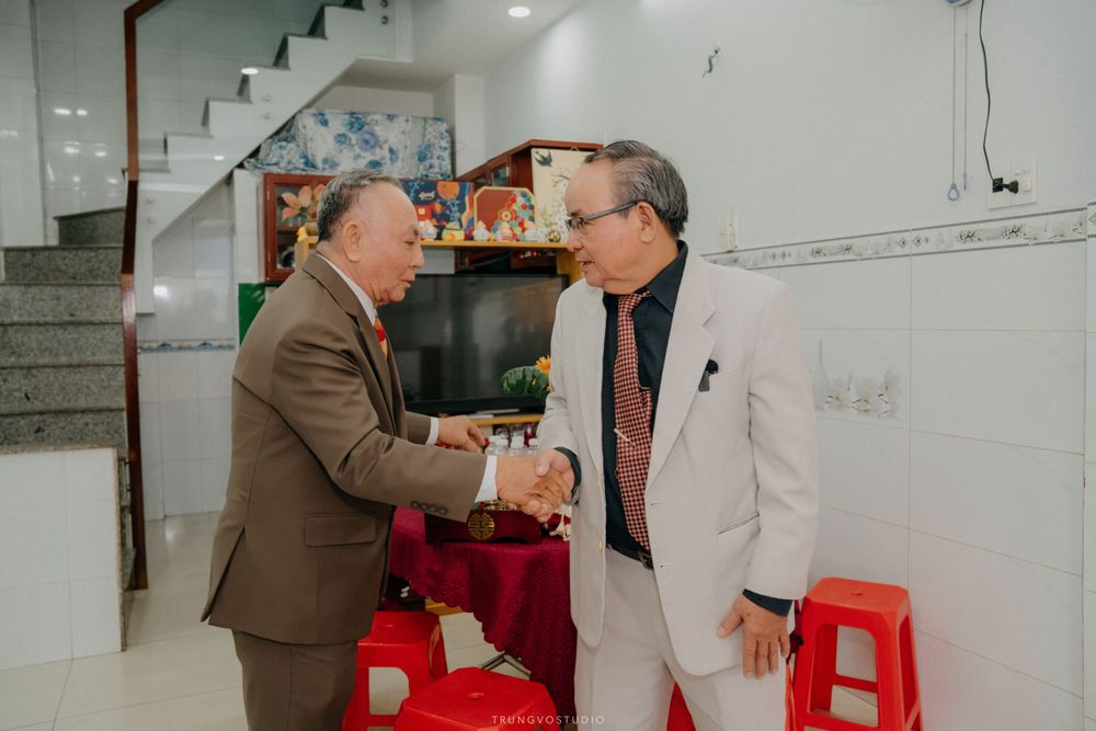 Two elderly men in formal attire shake hands warmly at a Vietnamese wedding celebration.