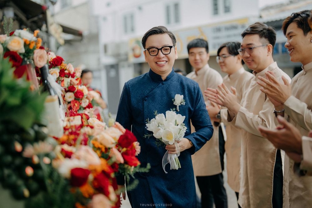 Smiling groom in blue ao dai holds white bouquet surrounded by applauding groomsmen.