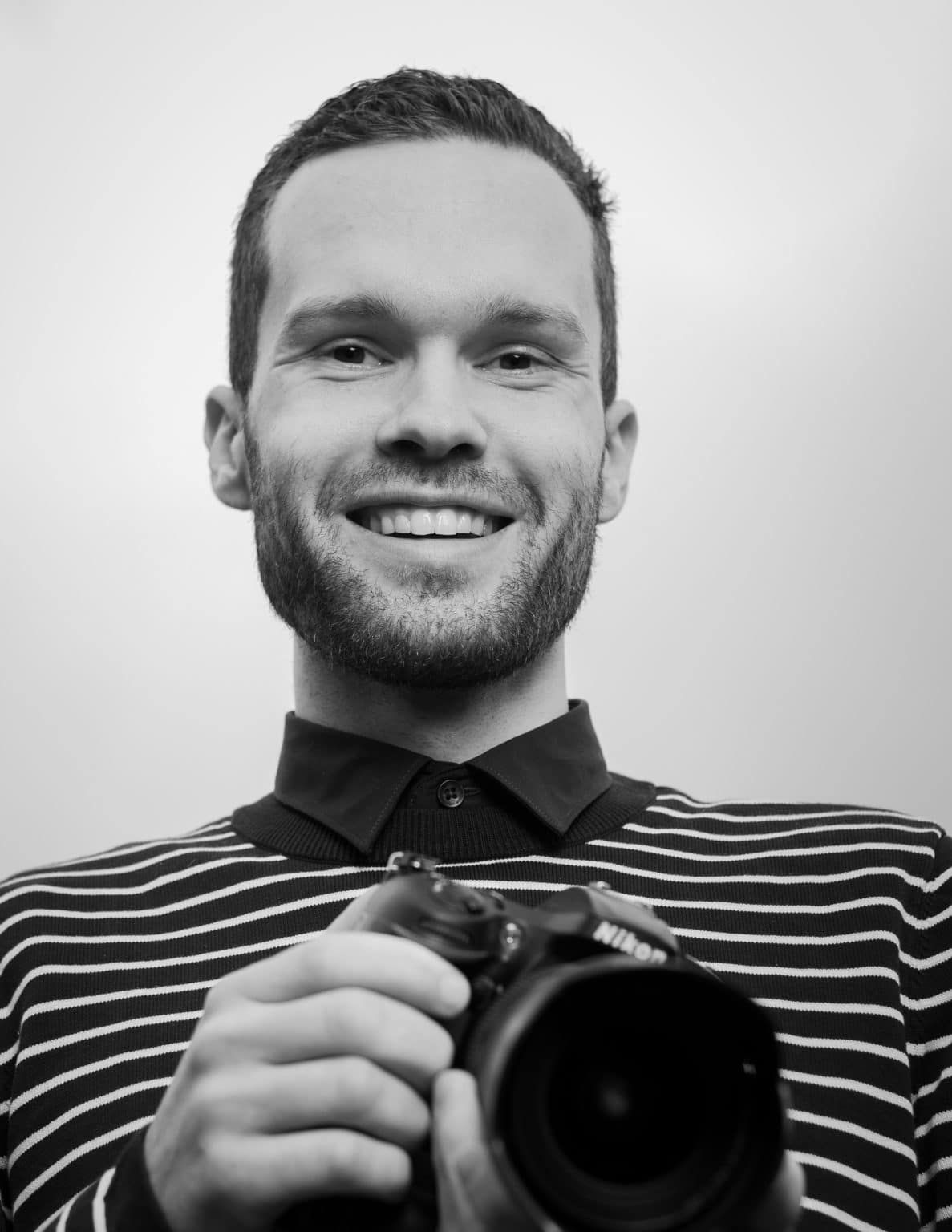 Photographer in black and white striped sweater smiles while holding professional camera against light background.