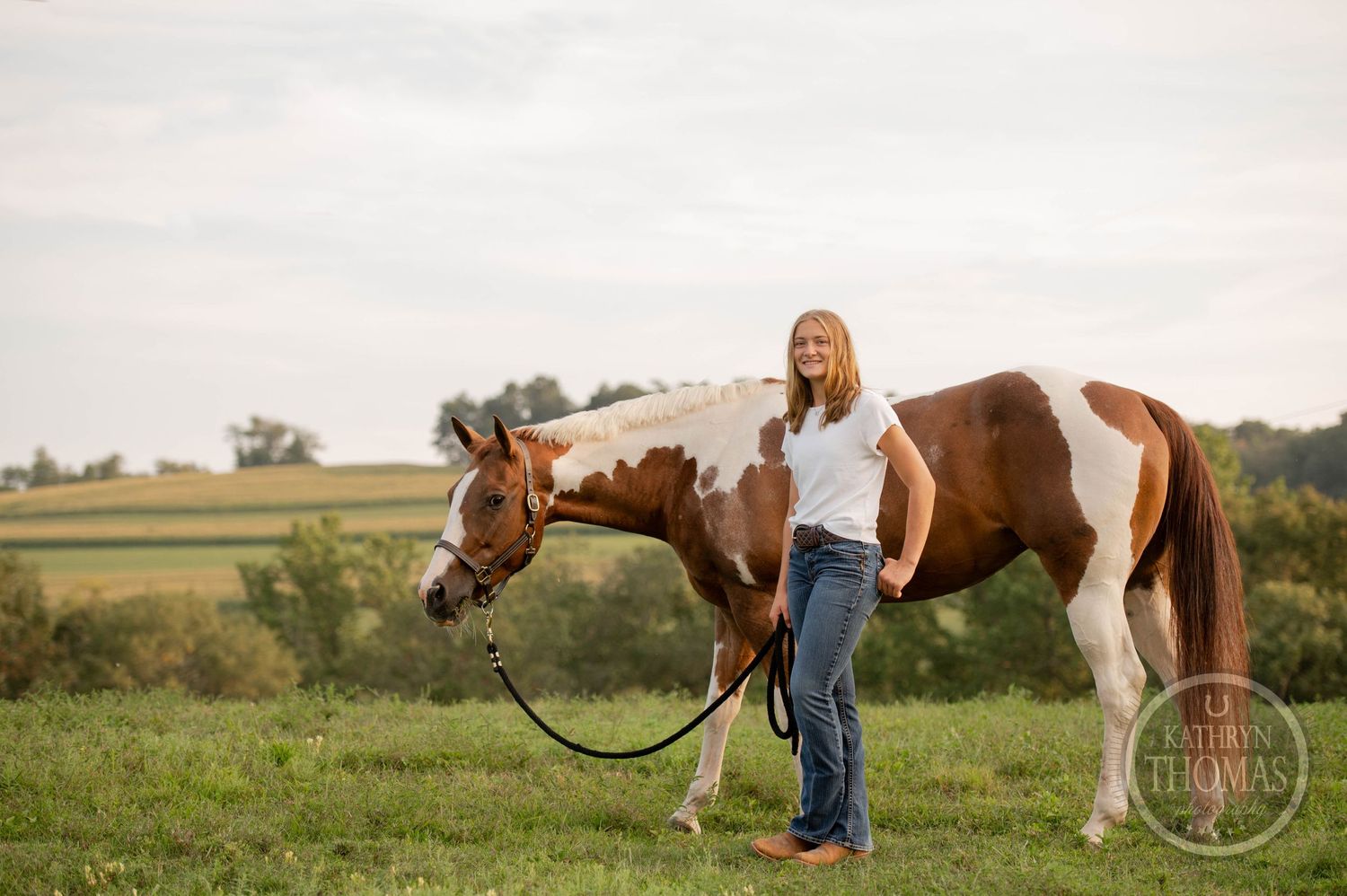 A brown and white paint horse stands in a grassy field at sunset with someone holding its lead rope.